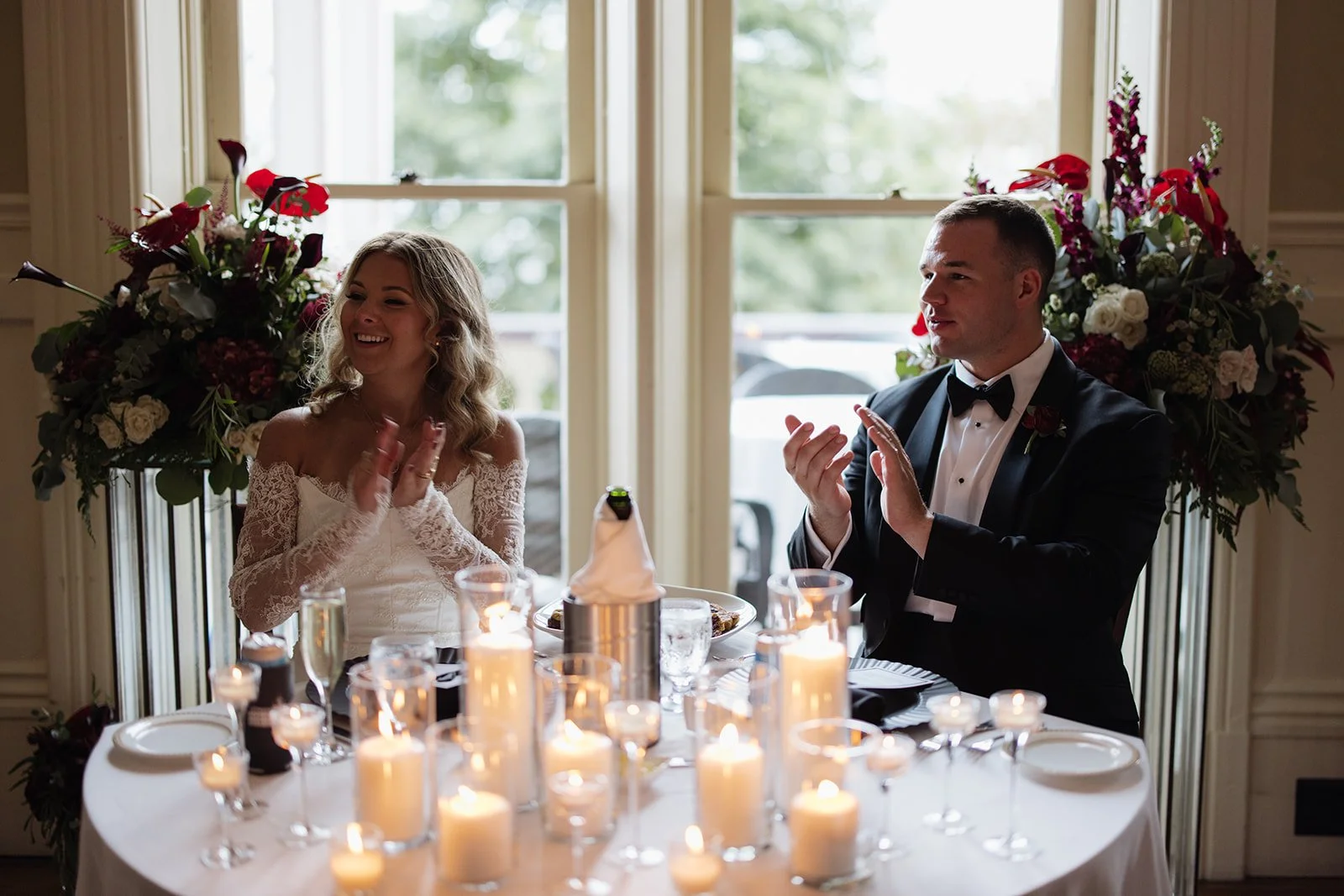 Bride and groom seated at a wedding reception table with lit candles and floral arrangements, clapping and smiling.