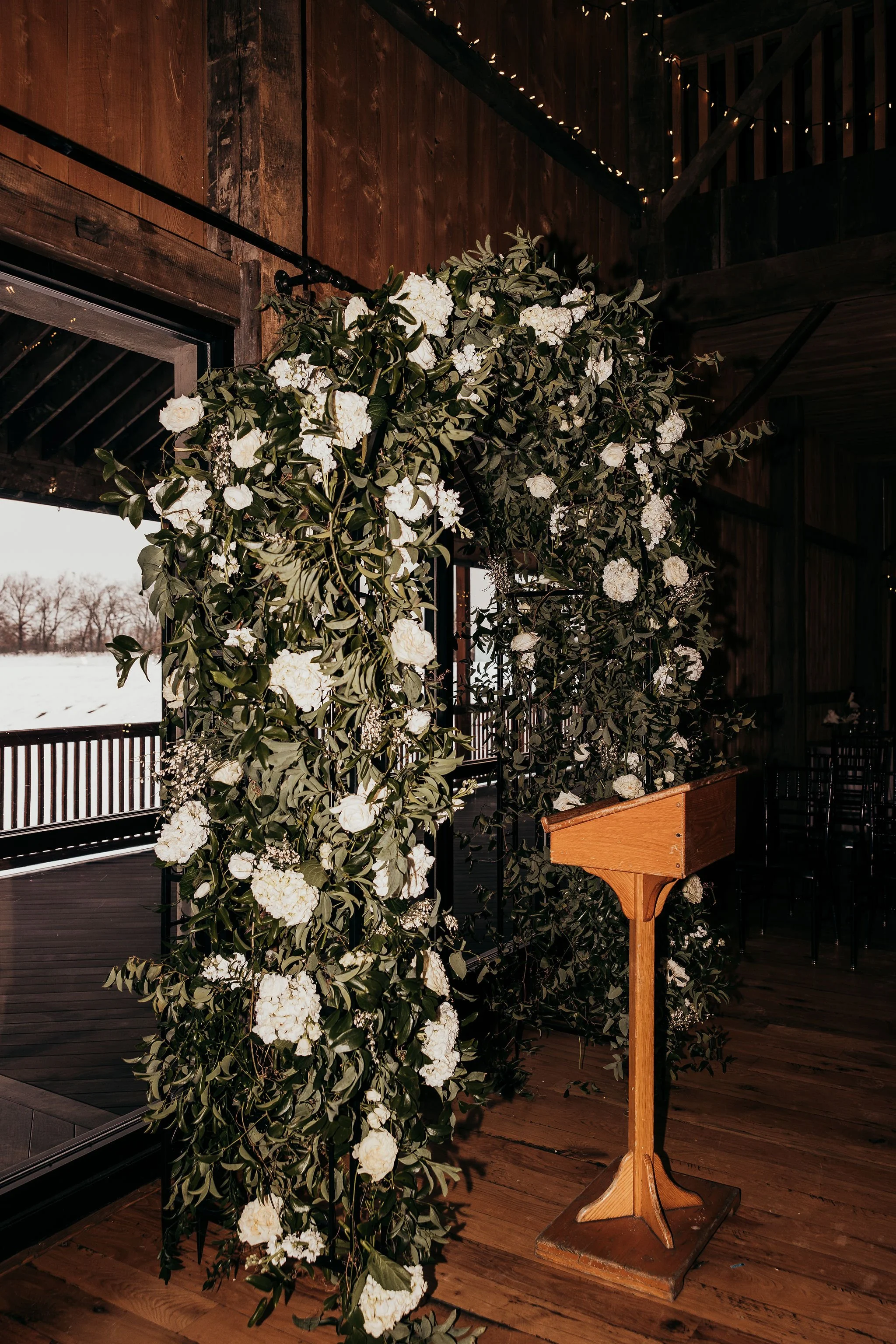 A wooden lectern stands in front of floral arrangements of white flowers and green foliage inside a rustic venue with wood-paneled walls and string lights. Outside, snow covers the landscape.