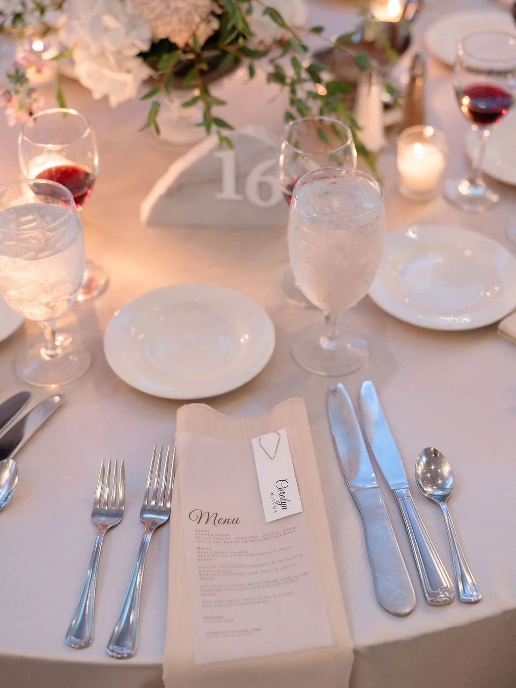 Elegant banquet table setting with white plates, silverware, wine glasses, water glasses, a menu, and a table number '16', decorated with floral arrangement and lit candles.