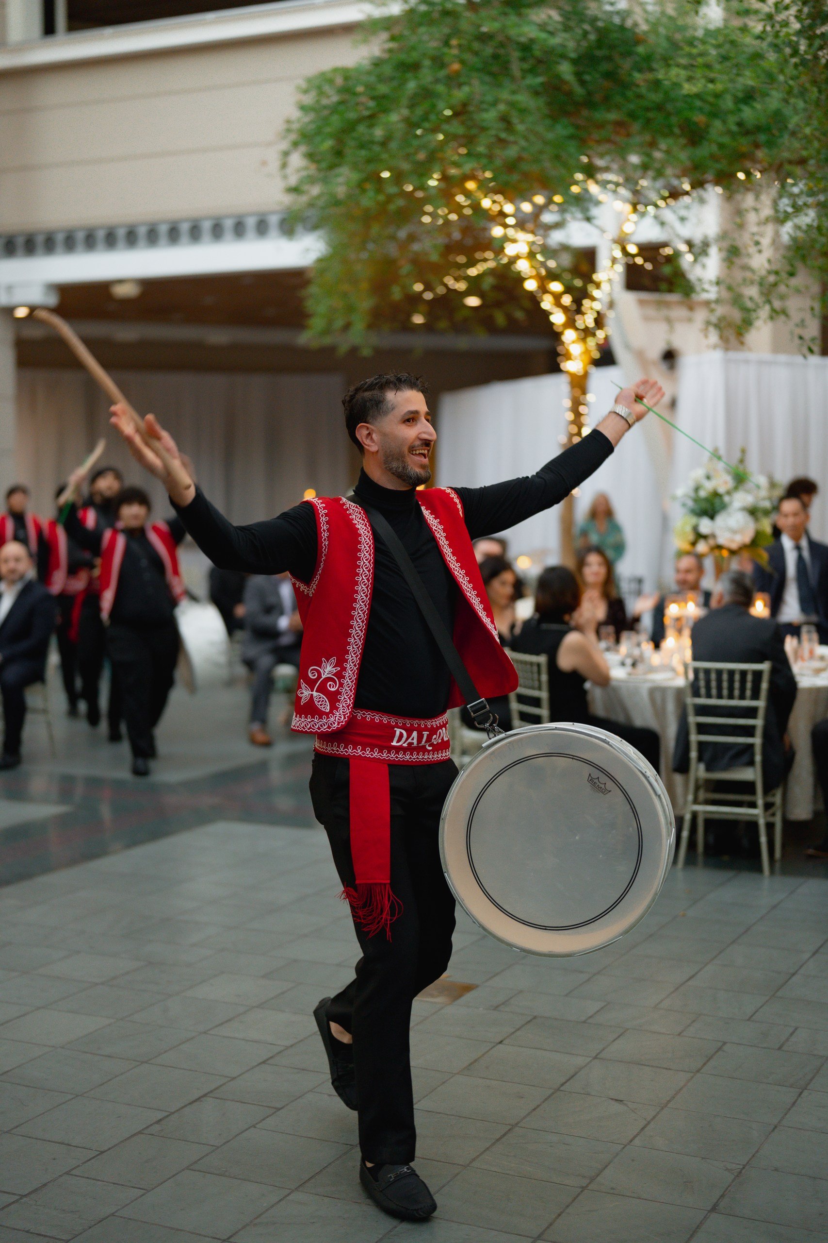 A man in black pants and shoes, wearing a red and black vest, is dancing and playing a large drum while holding a drumstick and a drumstick in his right hand. He is smiling and appears to be enjoying himself. The background shows a decorated ceiling 