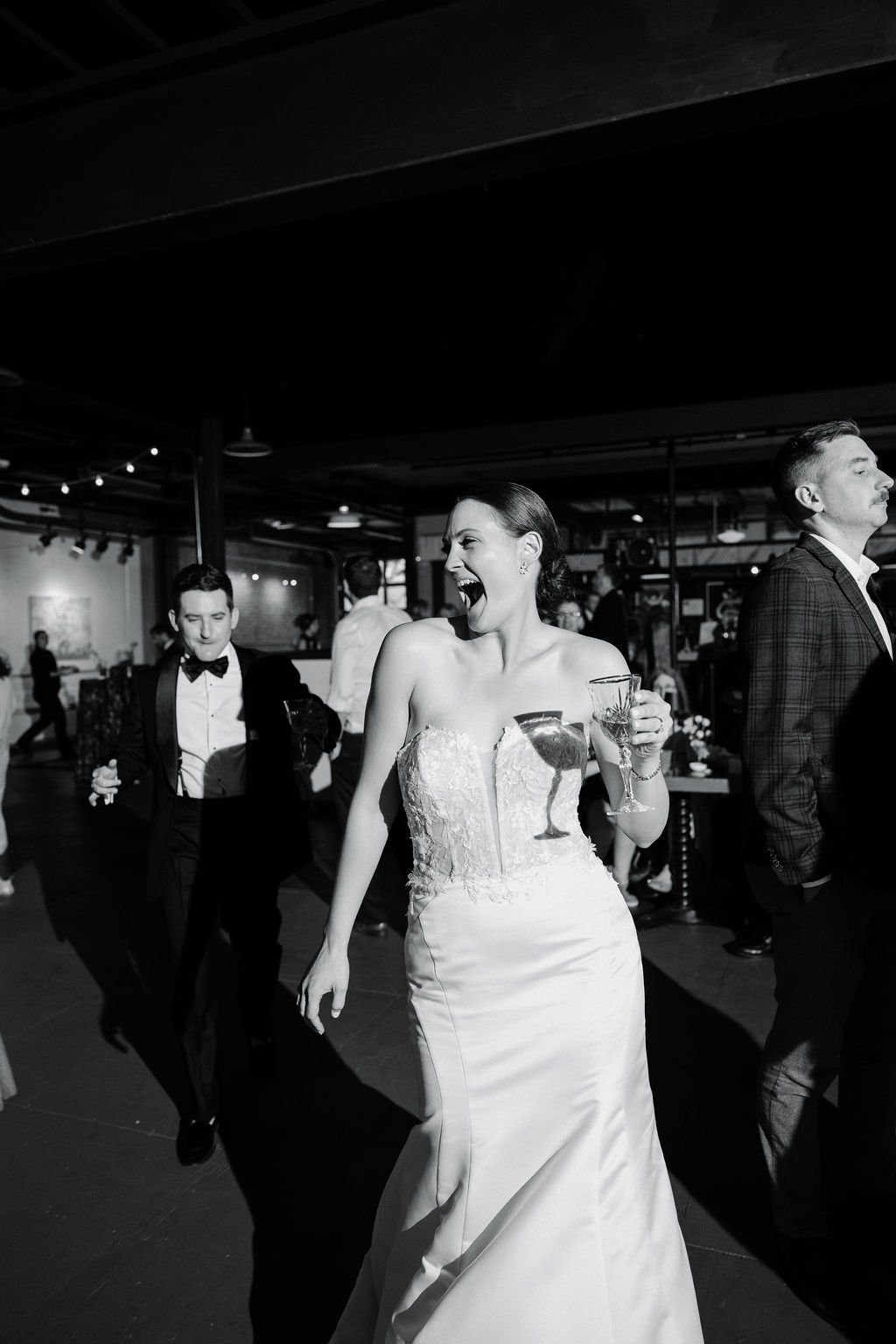 A woman in a wedding dress holding a glass, smiling and dancing at a wedding reception with guests in formal attire in the background.