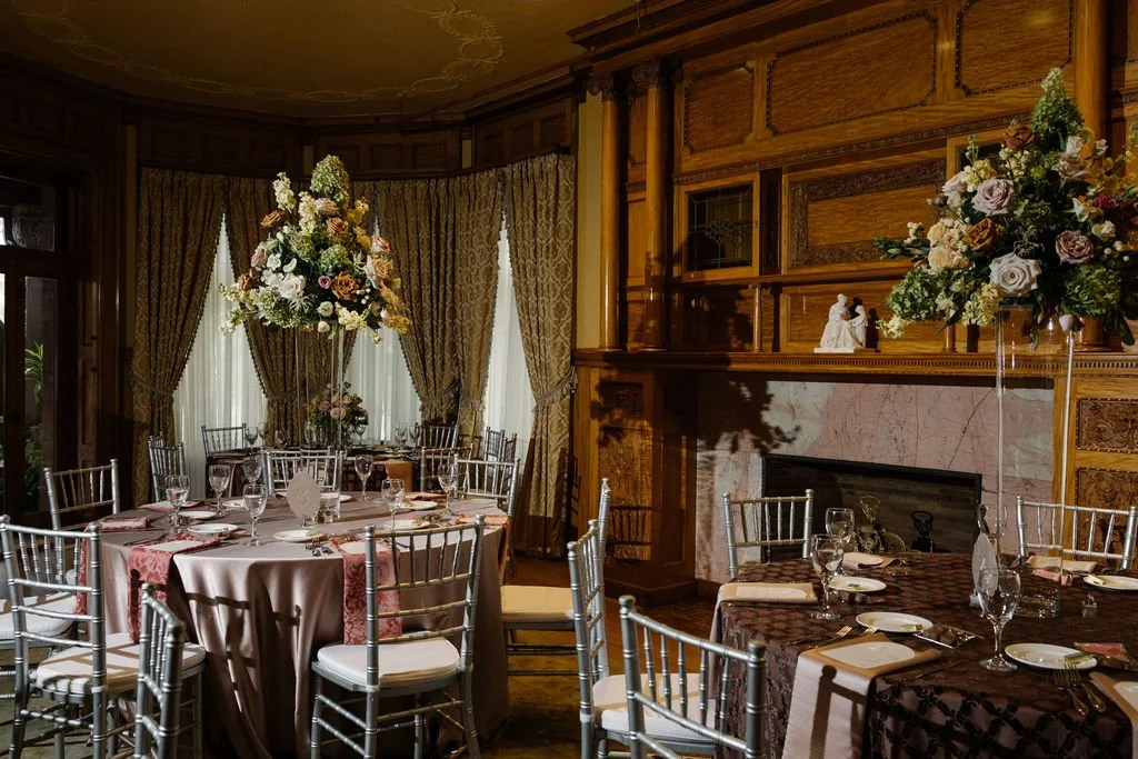 Elegant dining room with round tables, silver chairs, floral centerpieces, and a fireplace with wooden paneling.