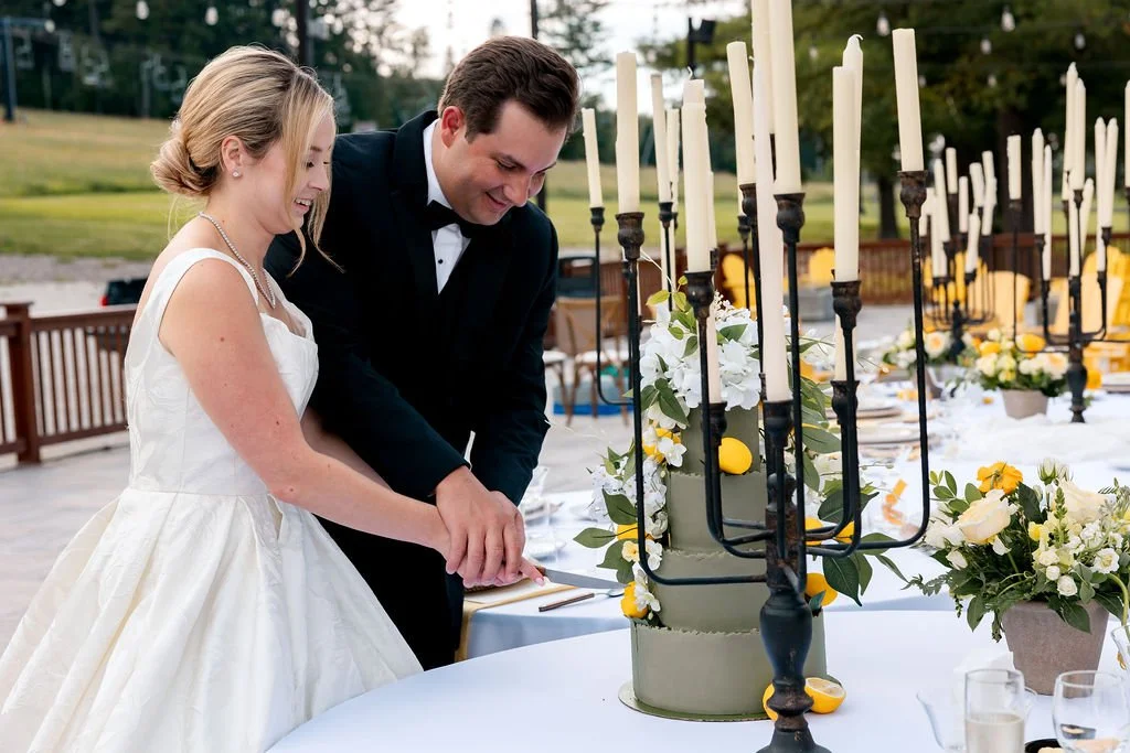 A bride and groom cutting their wedding cake at an outdoor reception, with tall candle holders and floral arrangements on the table.