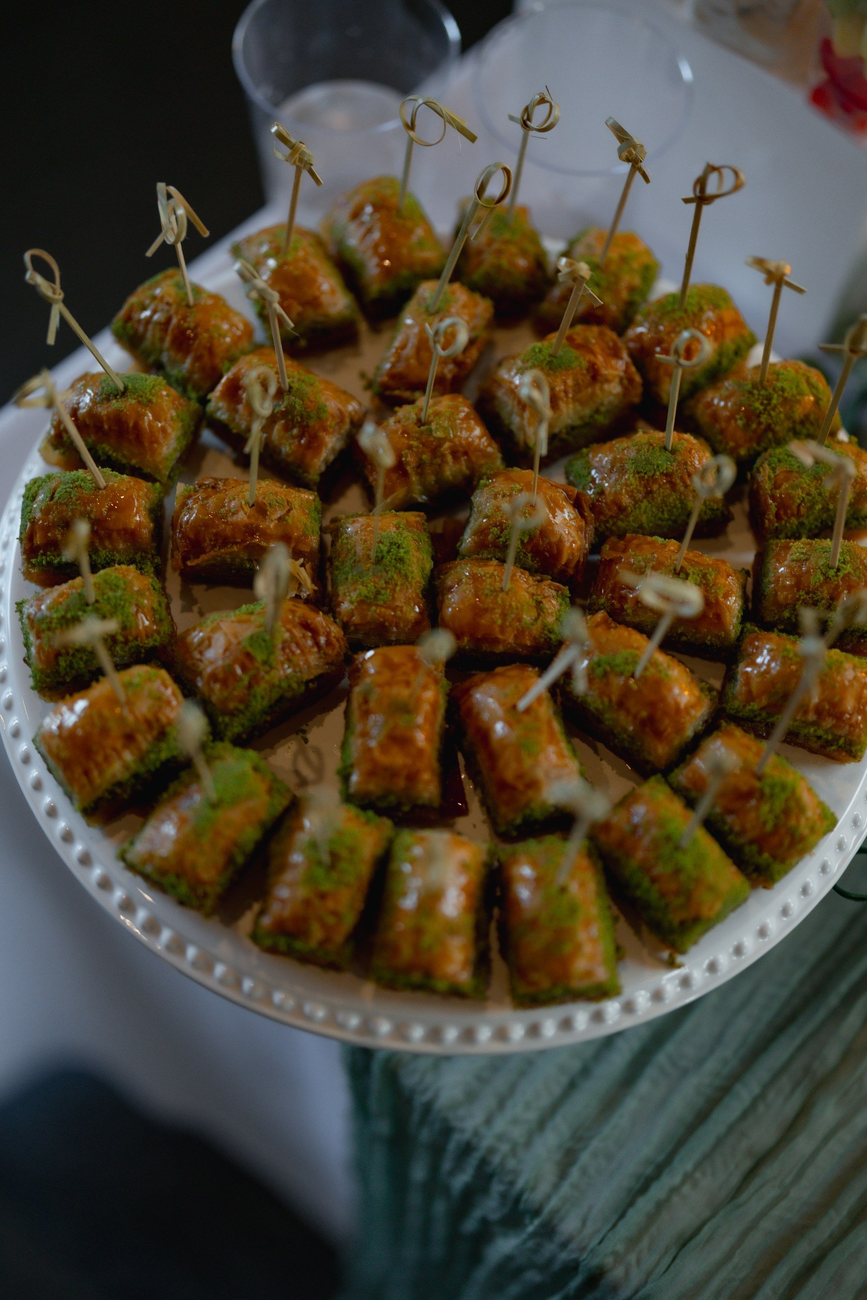 A white plate filled with small, square-shaped appetizers topped with a green herb seasoning and drizzled with a brown sauce. Each piece has a toothpick inserted for serving, and two clear plastic cups are visible in the background.