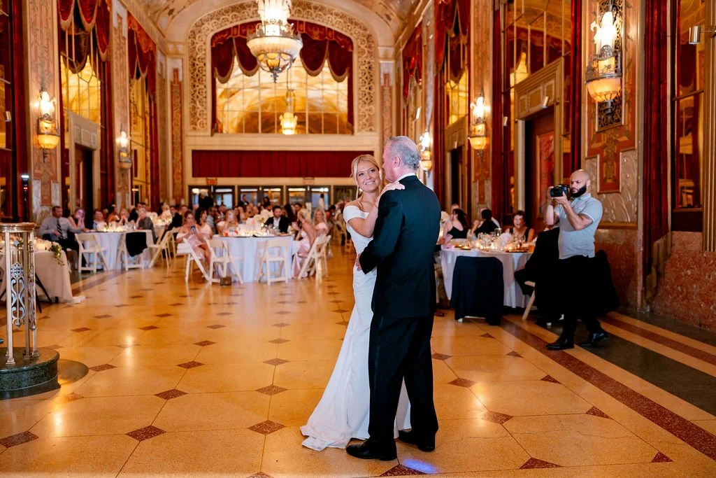 A bride and groom sharing a dance at their wedding reception in a grand, ornate ballroom with chandeliers and round tables filled with guests.