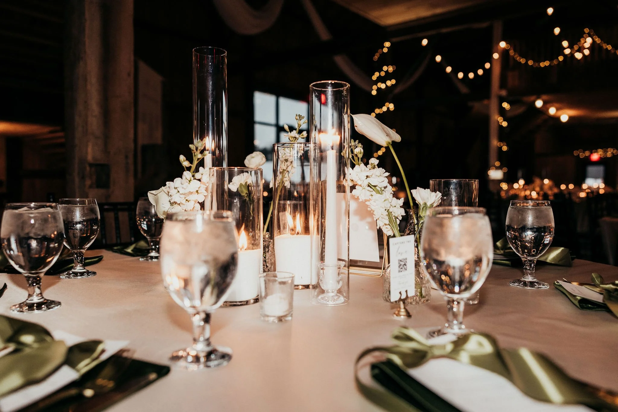 A decorated table with white floral centerpieces, tall glass candle holders, and wine glasses in a dimly lit room with string lights.
