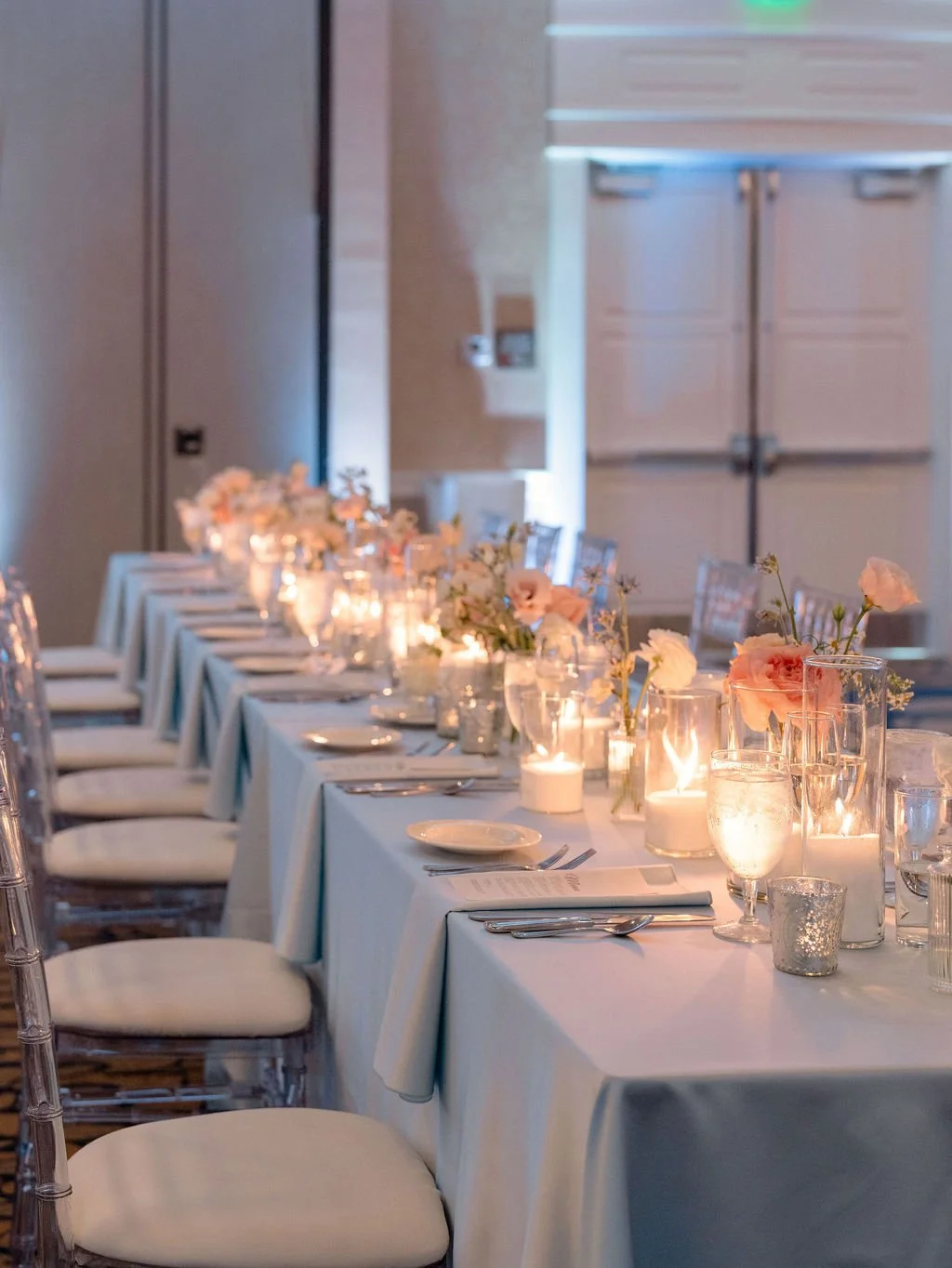 A long banquet table set for an event, decorated with candles, pink flowers, white tablecloth, and place settings with plates, silverware, and glasses.