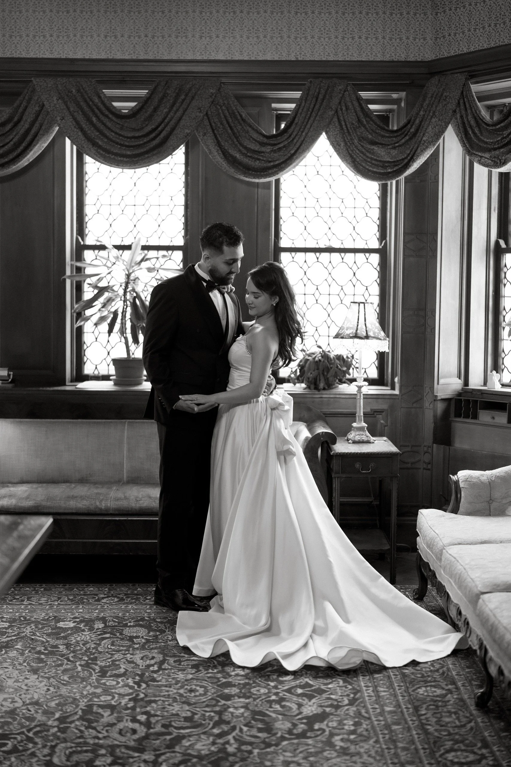 A black and white photo of a couple in wedding attire standing in a room with high windows, drapes, lamps, and vintage furniture, sharing a dance.