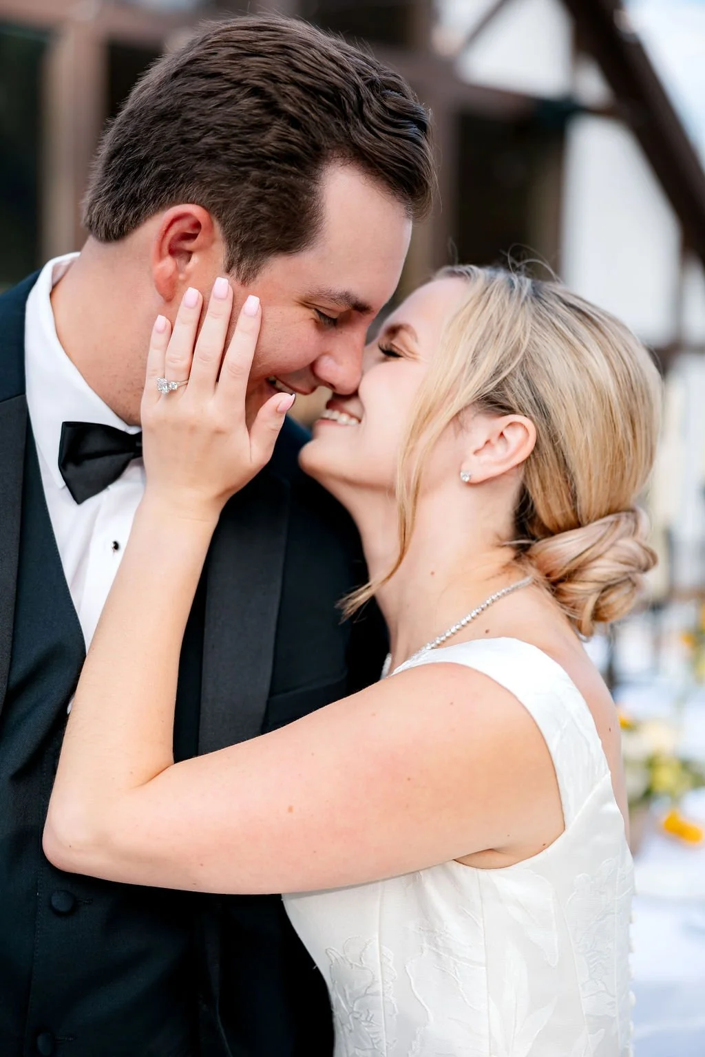 A newlywed couple sharing a close, intimate moment, smiling with noses touching. The woman’s hand, with a wedding ring, rests on the man’s face. The man is wearing a tuxedo with a bow tie, and the woman is in a white wedding dress with jewelry, both 