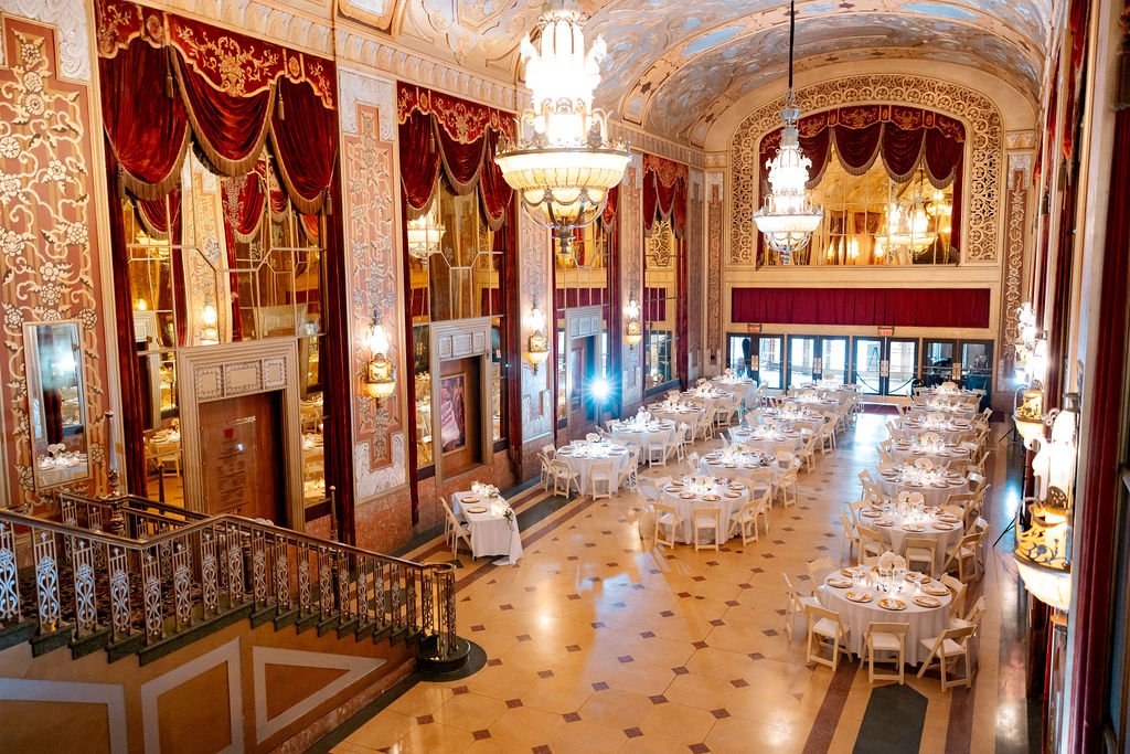 Elegant banquet hall with high, ornate ceilings, ornate chandeliers, large red draped curtains, and multiple round tables set for dining.