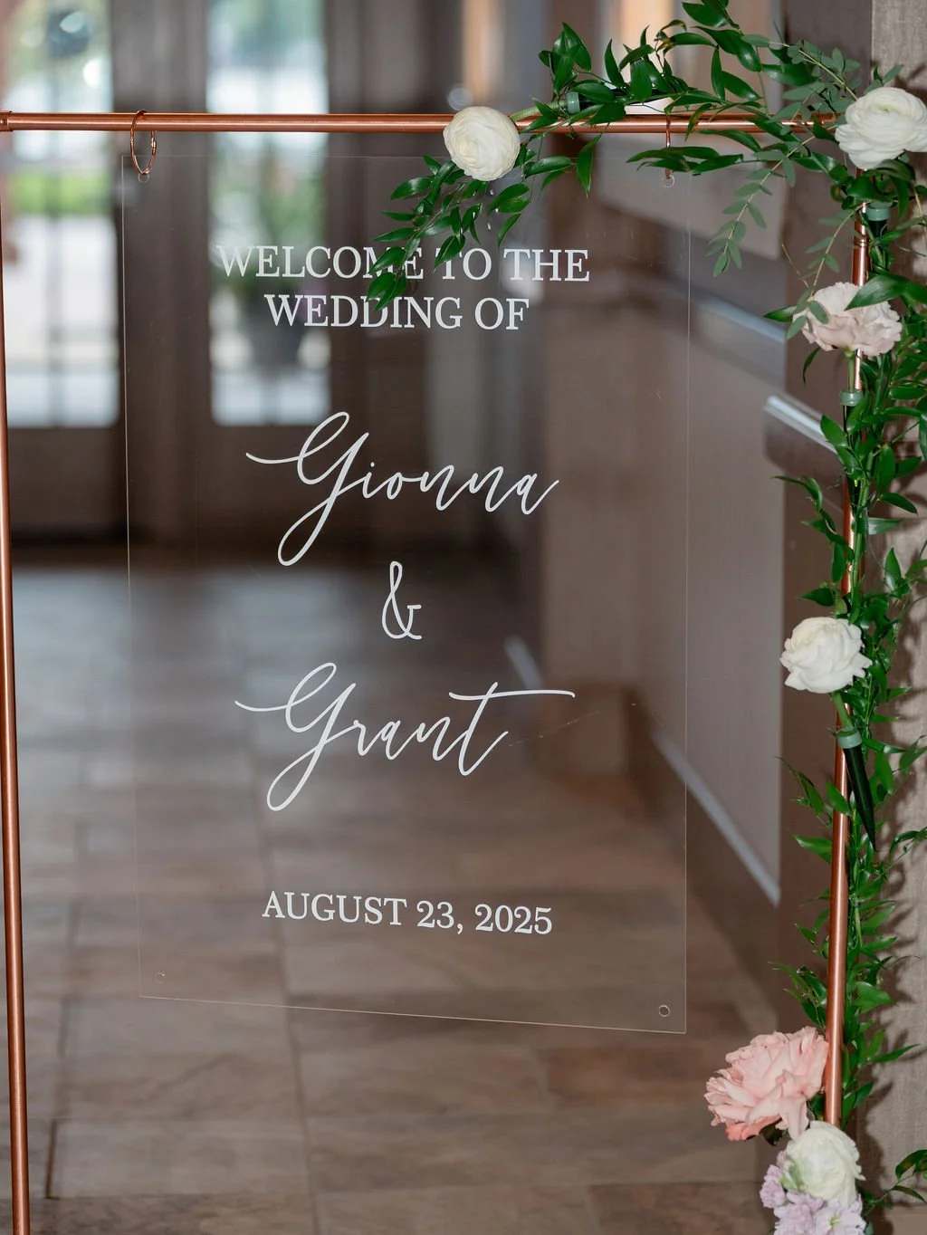 Wedding welcome sign decorated with green leaves and white and pink flowers, displaying the names Gioda and Grant and the date August 23, 2025.