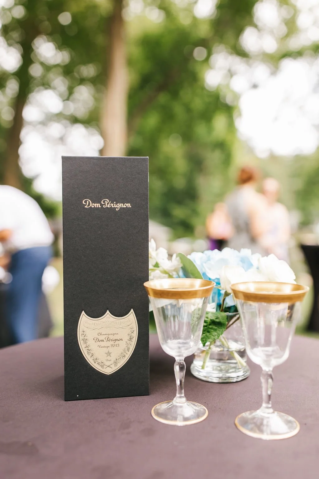 A black menu card with gold lettering, two champagne glasses with gold rims, and a floral centerpiece on a black tablecloth at an outdoor event with blurred people and trees in the background.