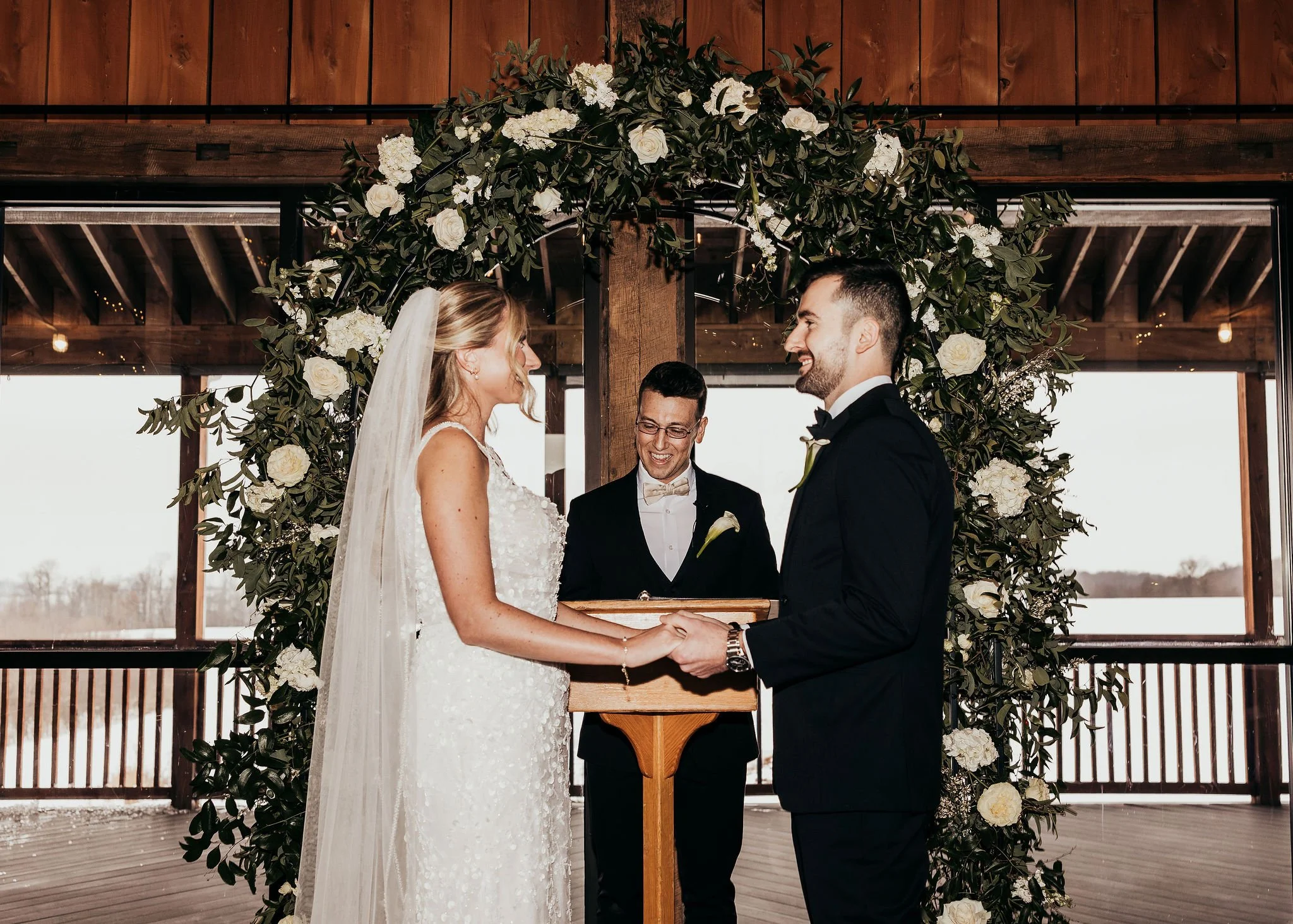 A bride and groom exchange vows during a wedding ceremony under a floral arch with greenery and white flowers, indoors with wooden accents and large windows.