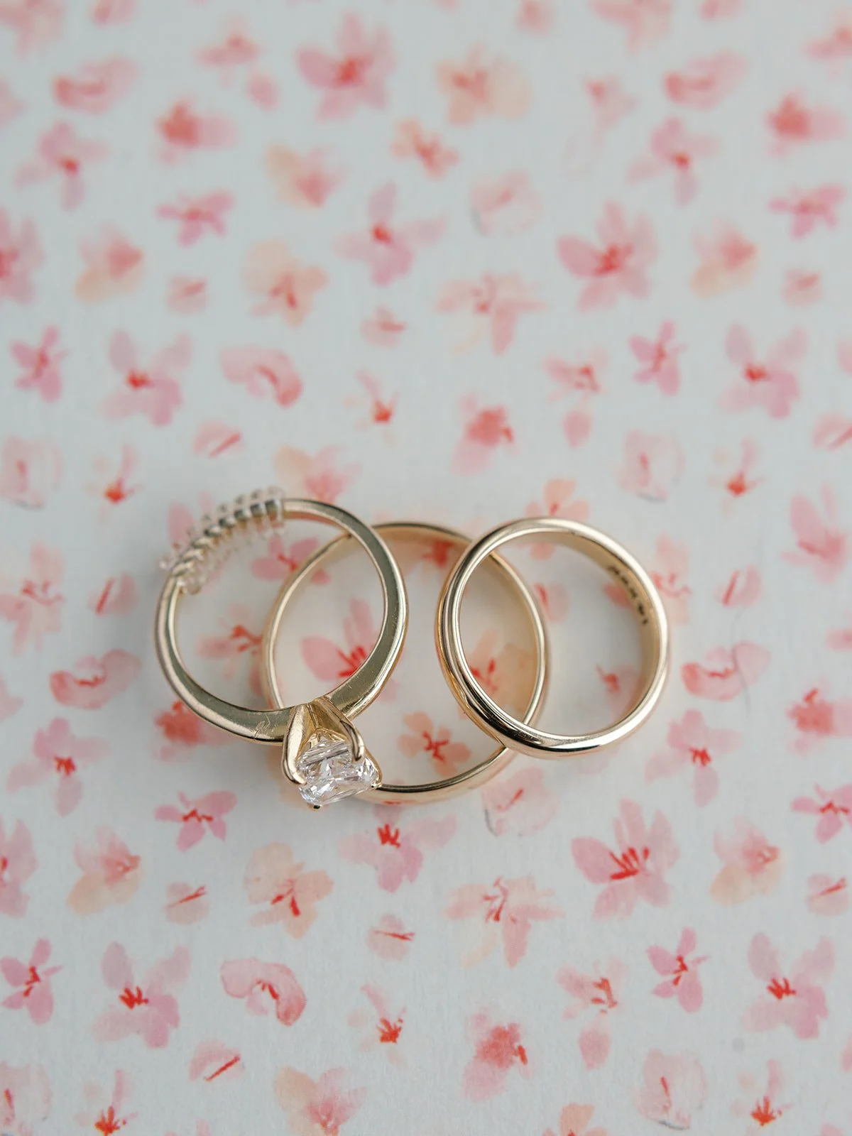 Three rings placed on a pink floral background, including a diamond engagement ring and two wedding bands.