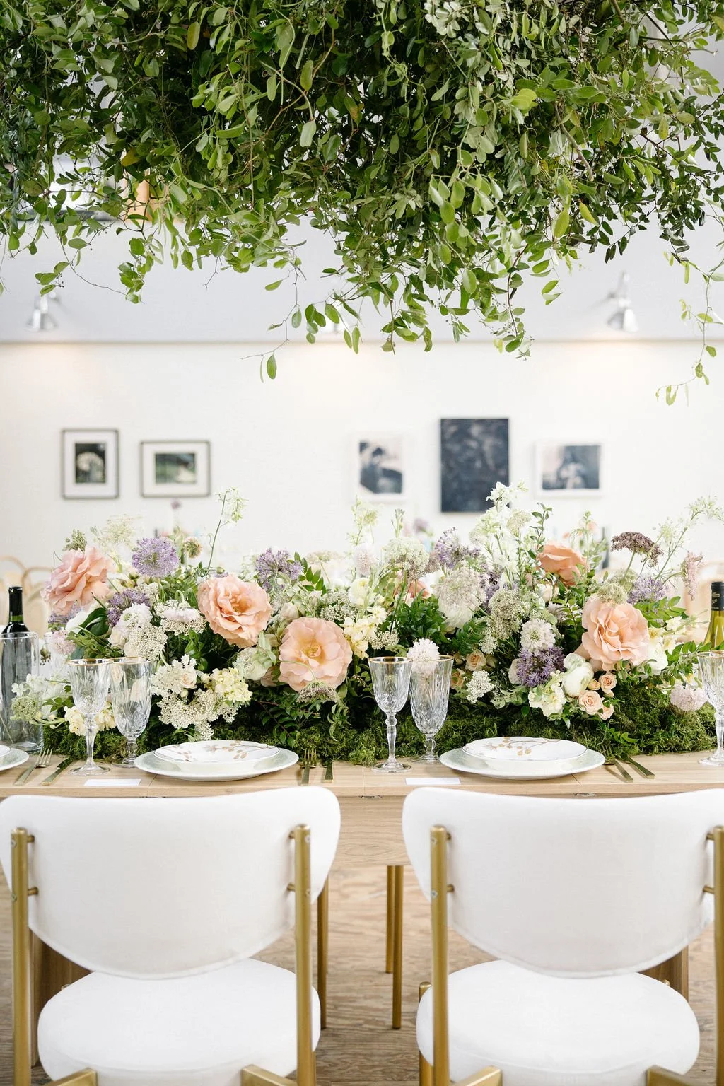 Elegant dining table decorated with a lush floral arrangement featuring pink and white flowers, surrounded by white chairs with gold accents, with a ceiling installation of greenery overhead in a well-lit room.