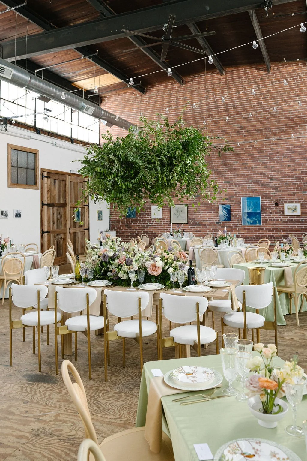 A decorated event space with round tables, floral centerpieces, and white and pastel chairs, featuring an indoor brick wall and hanging greenery.