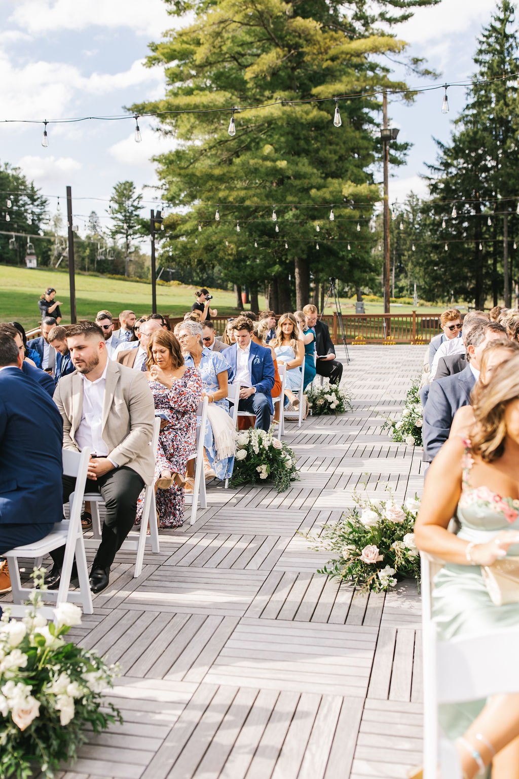 Guests seated outdoors at a wedding ceremony with floral arrangements and string lights, on a wooden deck with a scenic green landscape in the background.