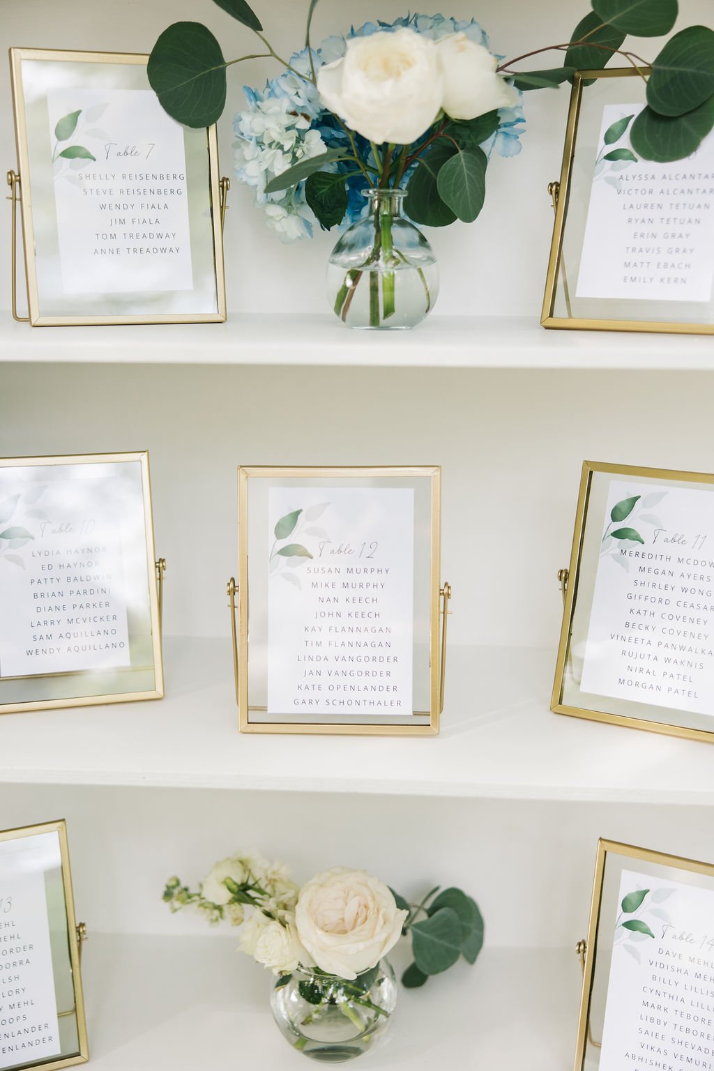 A display of elegant seating arrangement cards with gold frames and white paper with green leaf designs, small floral arrangements in clear vases, on a white surface.