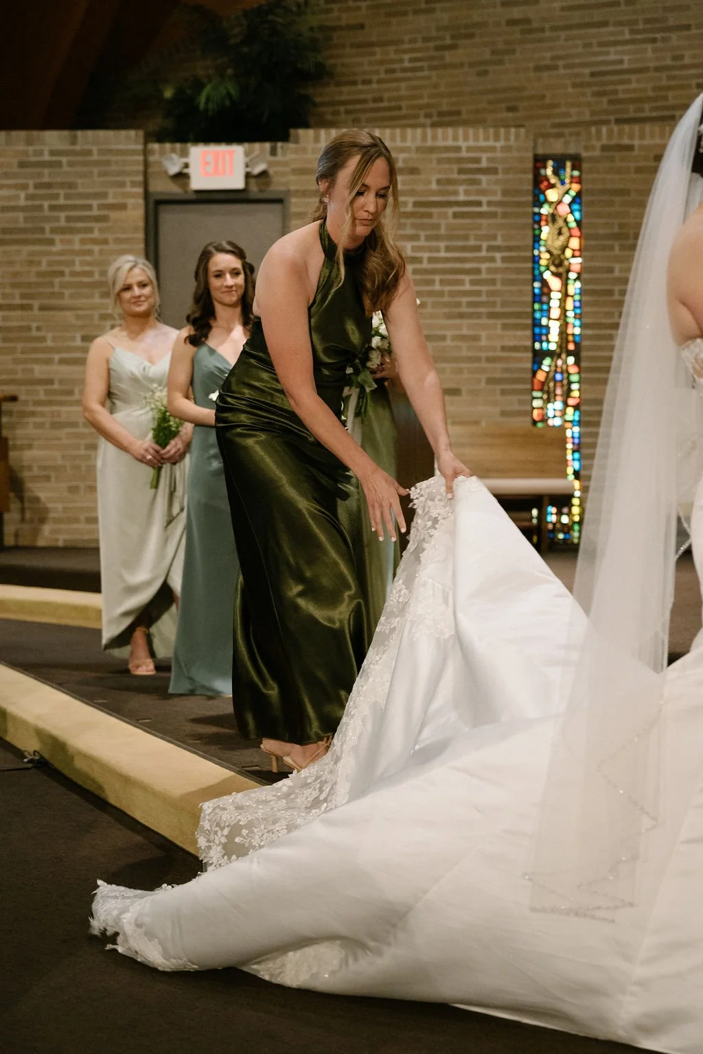 A woman in a shiny green dress is removing a wedding dress train in a church during a wedding ceremony, with two women in dresses and holding flowers standing in the background.
