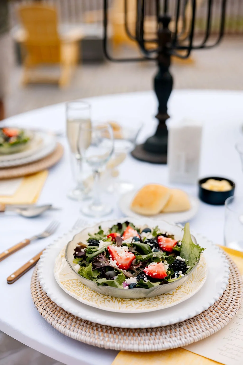 A table set for a meal with a fresh mixed green salad topped with strawberries and blackberries in a glass bowl, bread rolls, wine glasses, and a black lantern centerpiece.