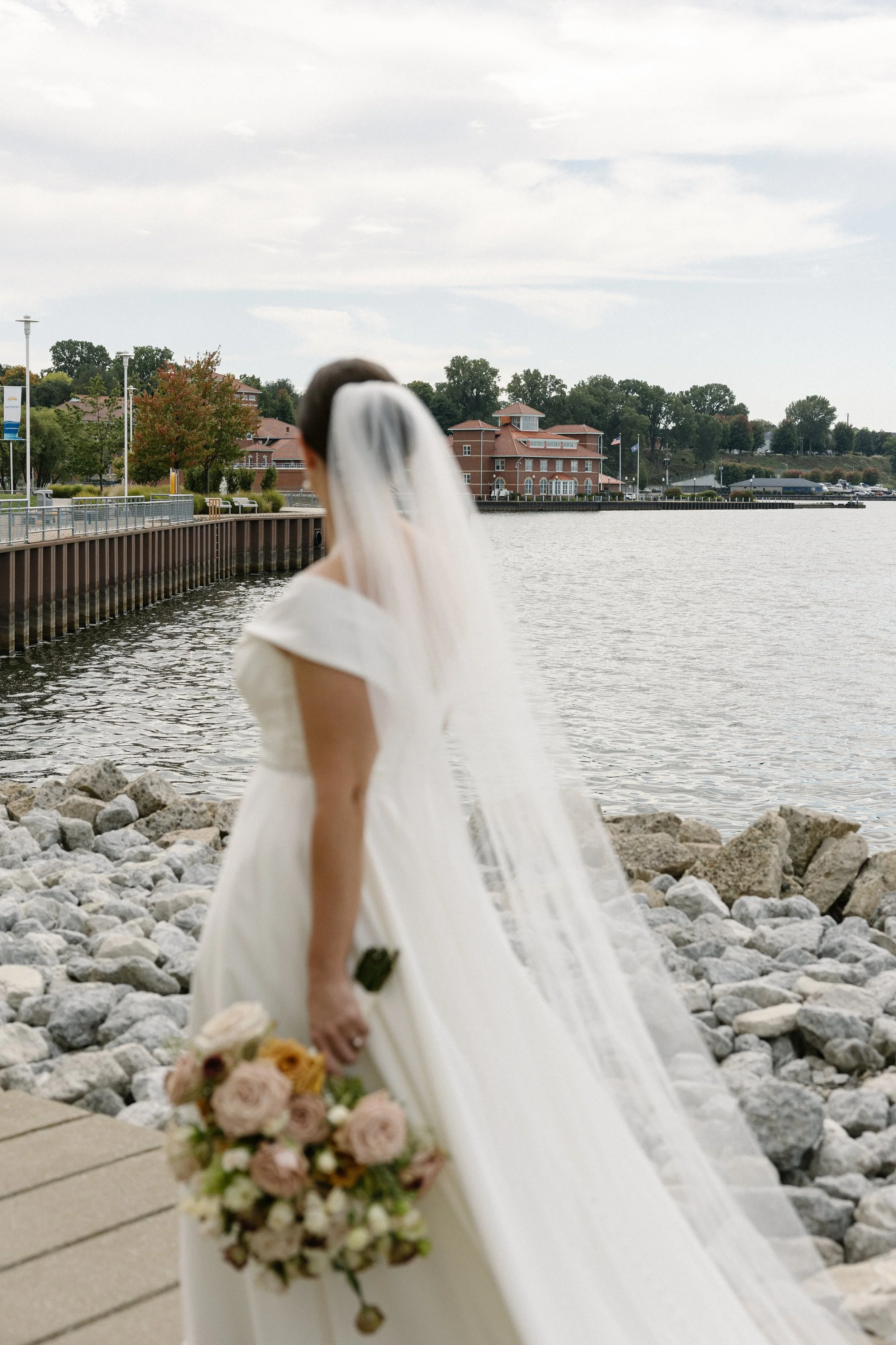 A bride holding a bouquet of pink, white, and peach flowers, standing on rocks by a body of water with buildings and trees in the background.