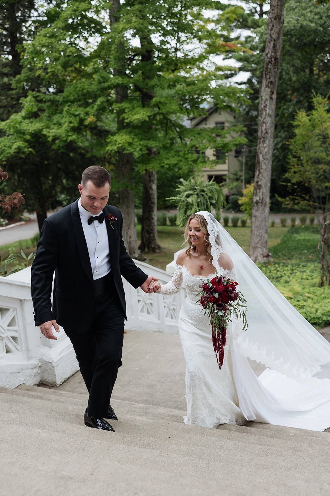 A bride and groom walking down the stairs outside on a wedding day, holding hands. The bride is wearing a white wedding gown with lace details and a long veil, holding a bouquet of red and dark purple flowers. The groom is dressed in a black tuxedo w