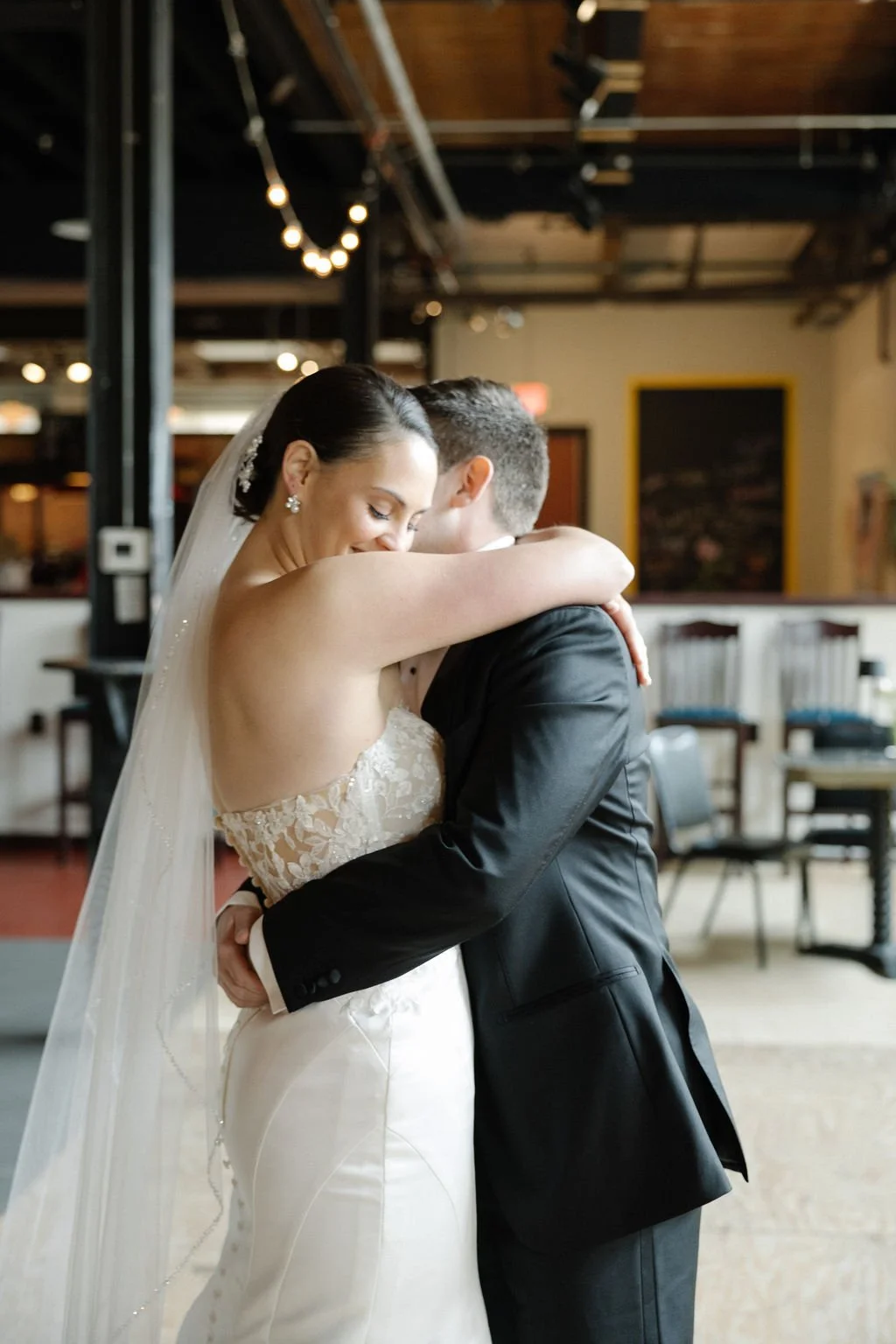 A bride and groom embrace during their wedding, indoor setting with warm lighting and blurred background.