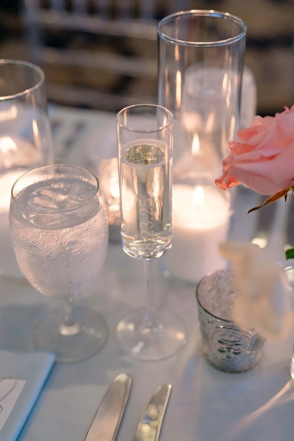 Set of elegant glassware including a water glass, a champagne flute with a clear beverage, a tall narrow glass, and a flower in a small vase on a table with lit candles.