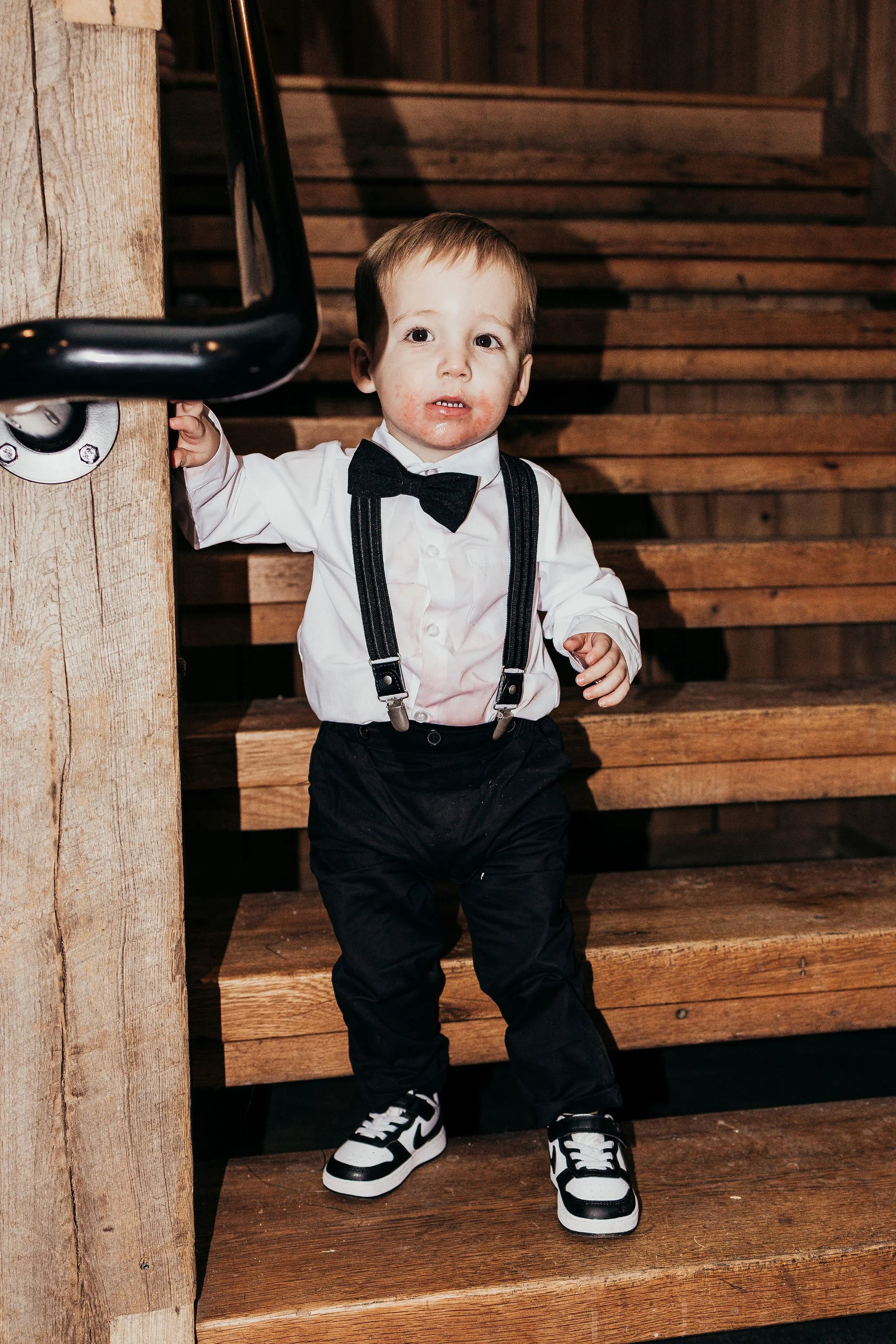 A young boy dressed in formal attire, including a white shirt with suspenders, black pants, and sneakers, stands on wooden stairs with a serious expression, touching a metal handrail.