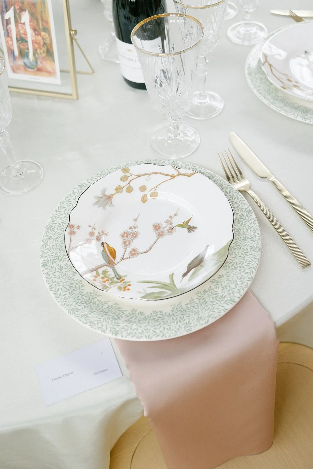 Elegant place setting with a decorative plate featuring a floral and bird design, a green leafy border, gold-rimmed glassware, and gold cutlery on a white tablecloth.