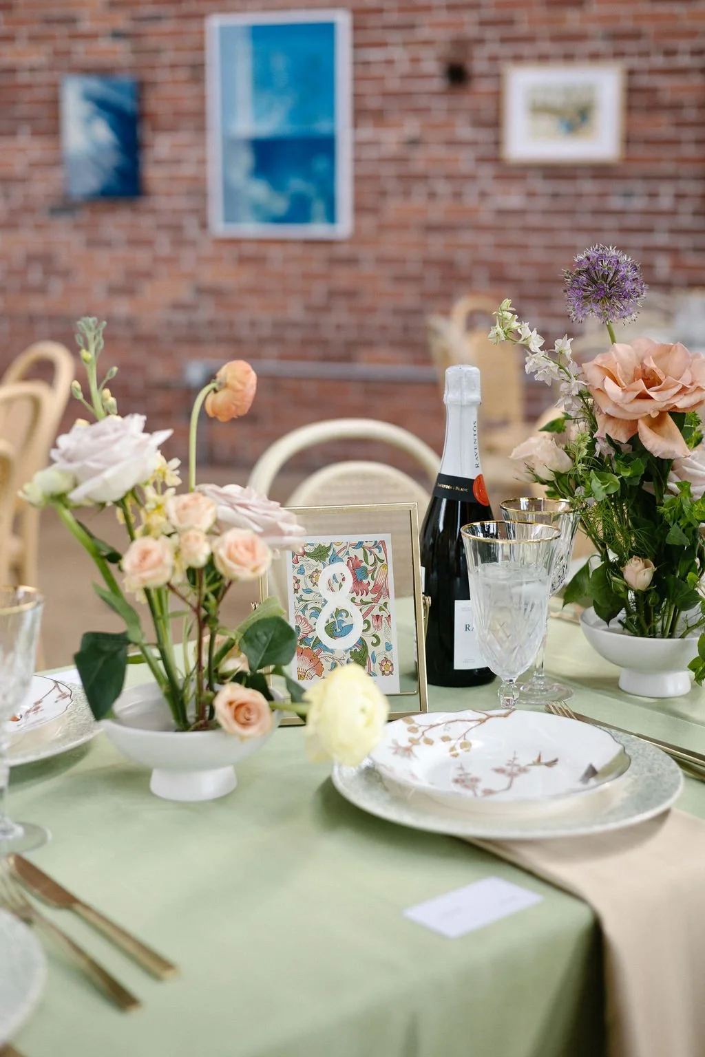 A decorated table with floral arrangements, a bottle of champagne, and elegant glassware, set against a brick wall with framed artwork.