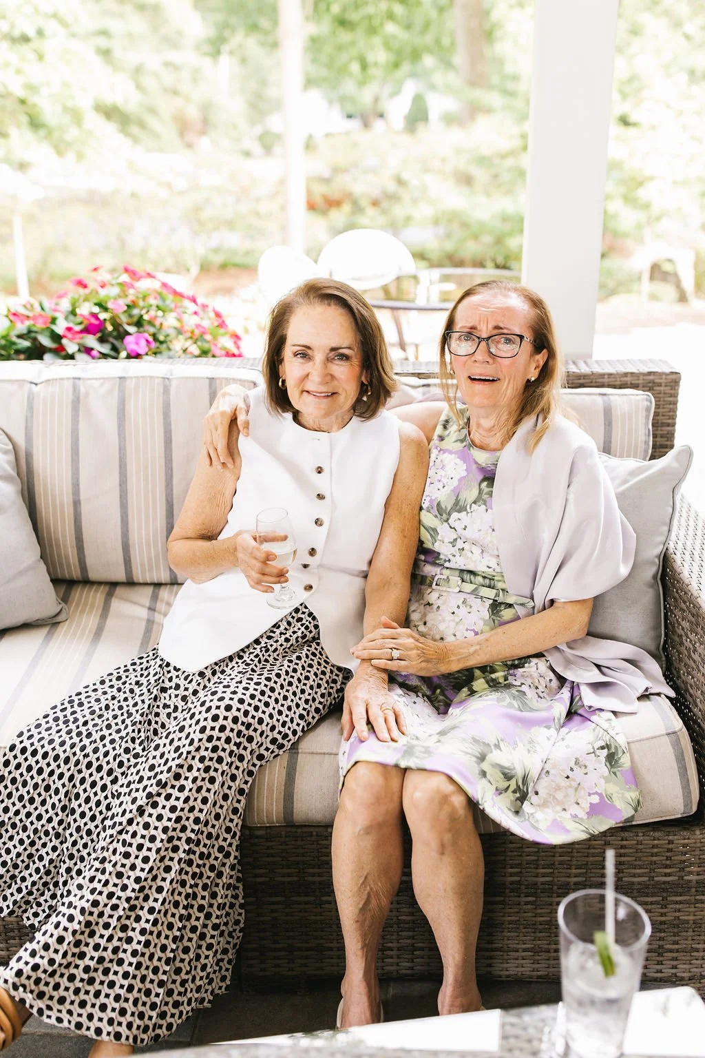 Two older women sitting on a patio sofa, smiling and holding drinks, with a background of greenery and flowers.