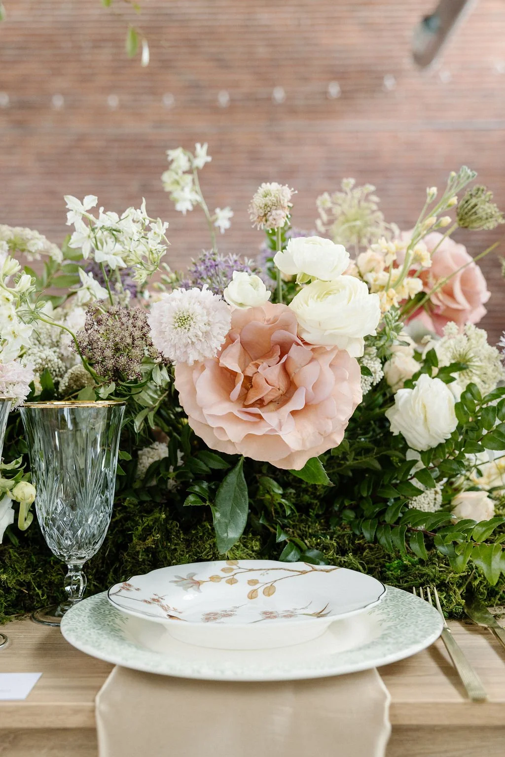 A floral centerpiece with pink, white, and purple flowers on a table set with elegant china, a glass, and gold-toned cutlery, against a wooden backdrop.