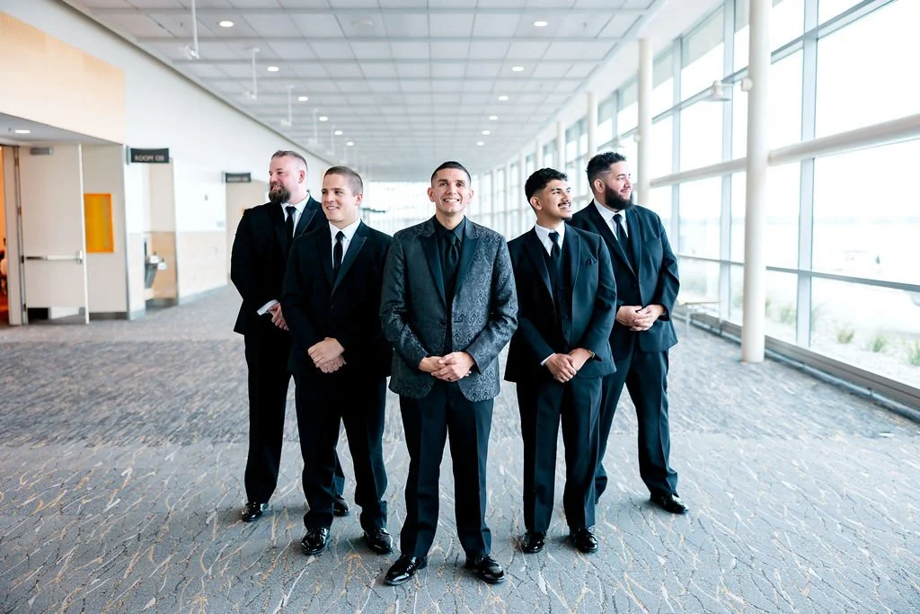 Five men in suits standing in a bright, modern hallway with large windows, smiling and looking in different directions.