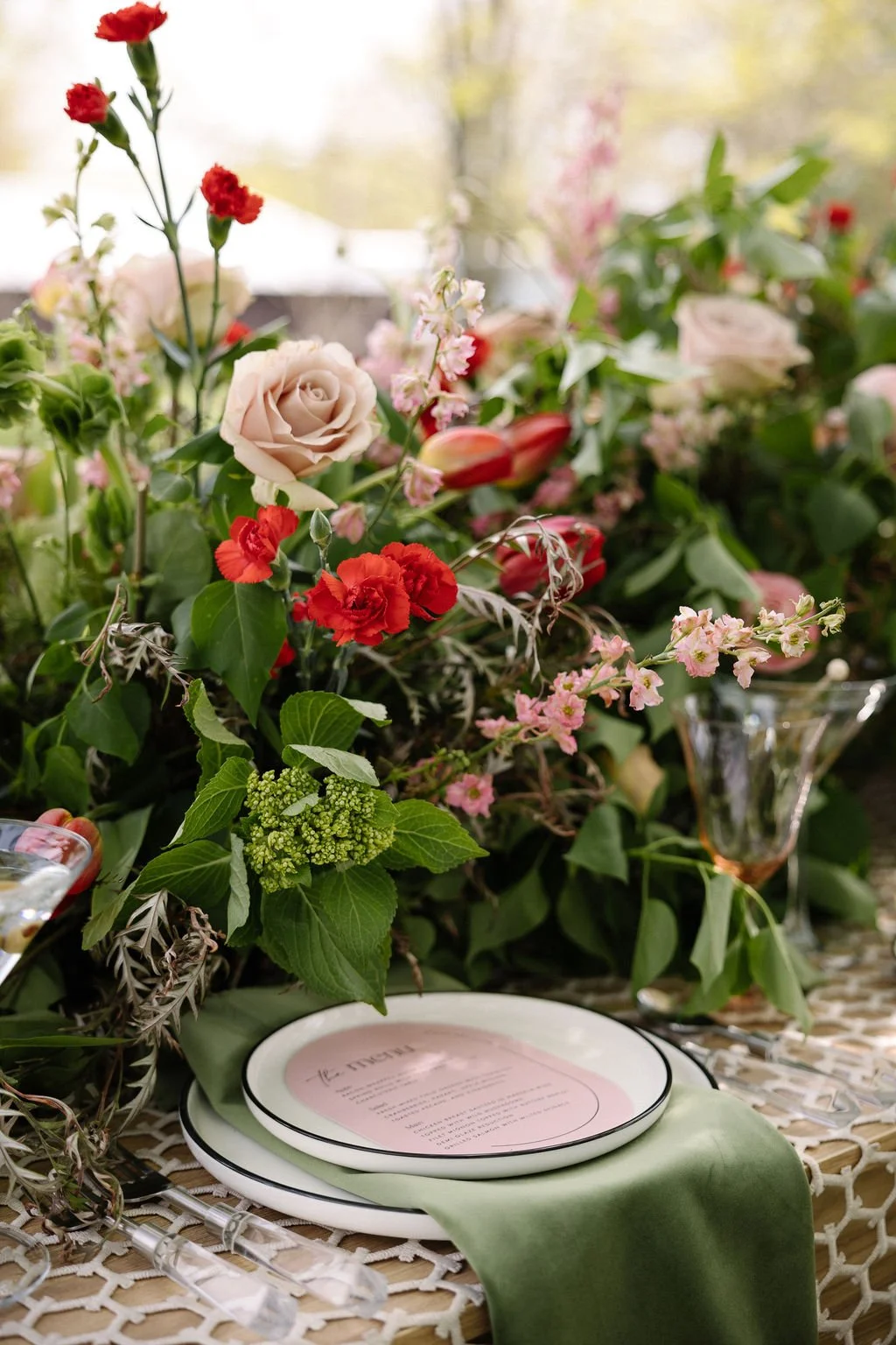 A floral arrangement with pink, red, and white flowers, including roses and assorted greenery, placed on a table with plates, a green napkin, and glassware, set in a well-lit environment.
