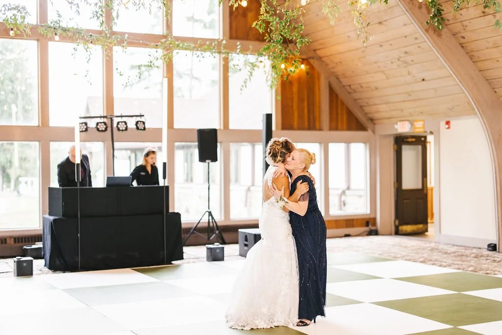 A bride and an older woman sharing a dance at a wedding reception in a spacious, rustic venue with large windows and wooden interior.