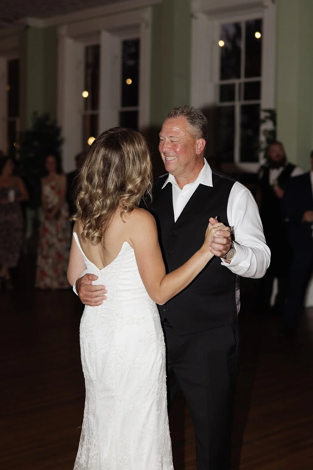 A couple dancing at a wedding reception, the woman in a white lace wedding dress and the man in a black vest and white shirt, smiling.