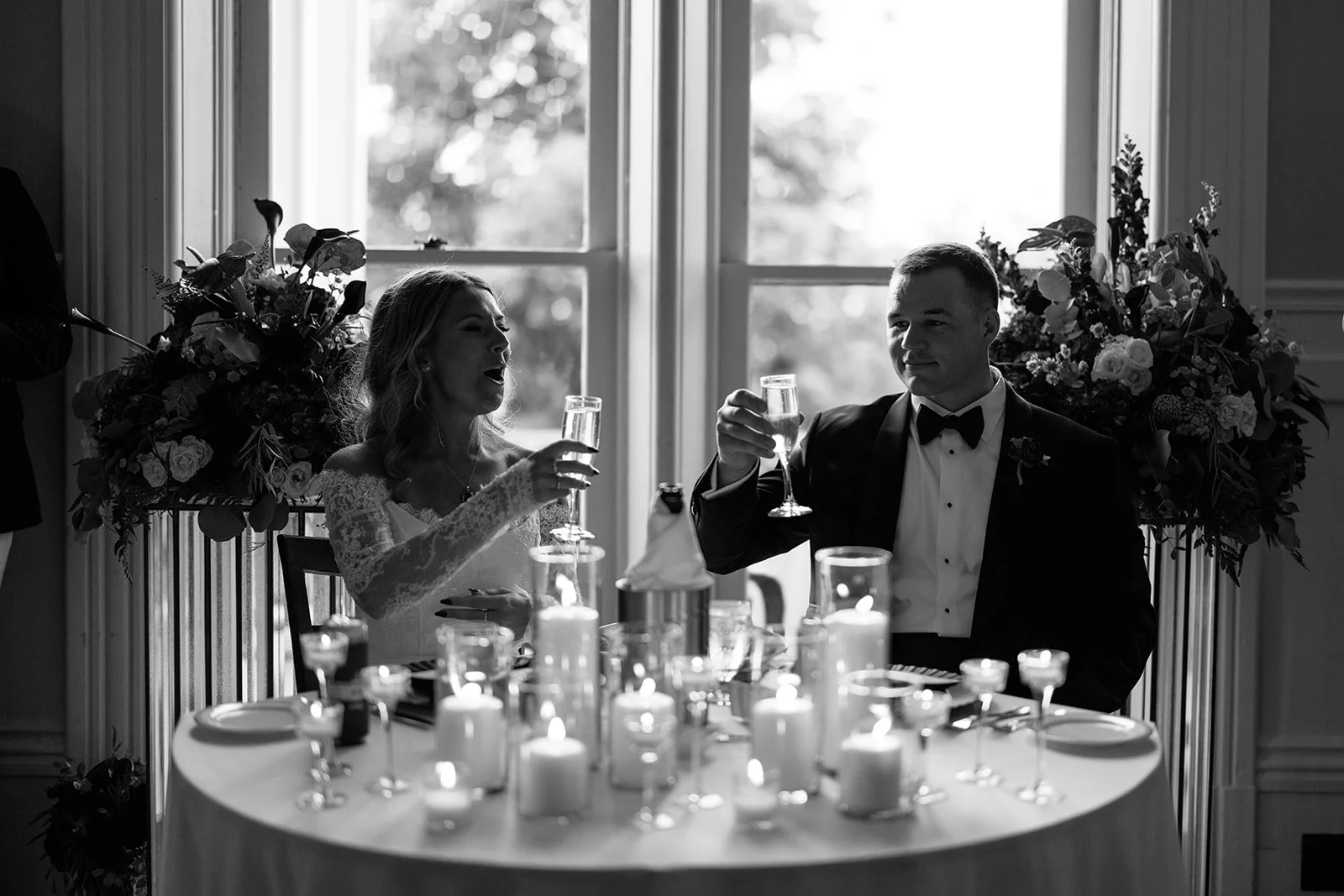 A black-and-white photo of a bride and groom celebrating at their wedding reception, raising champagne glasses near a decorated table with candles, floral arrangements, and elegant decor.