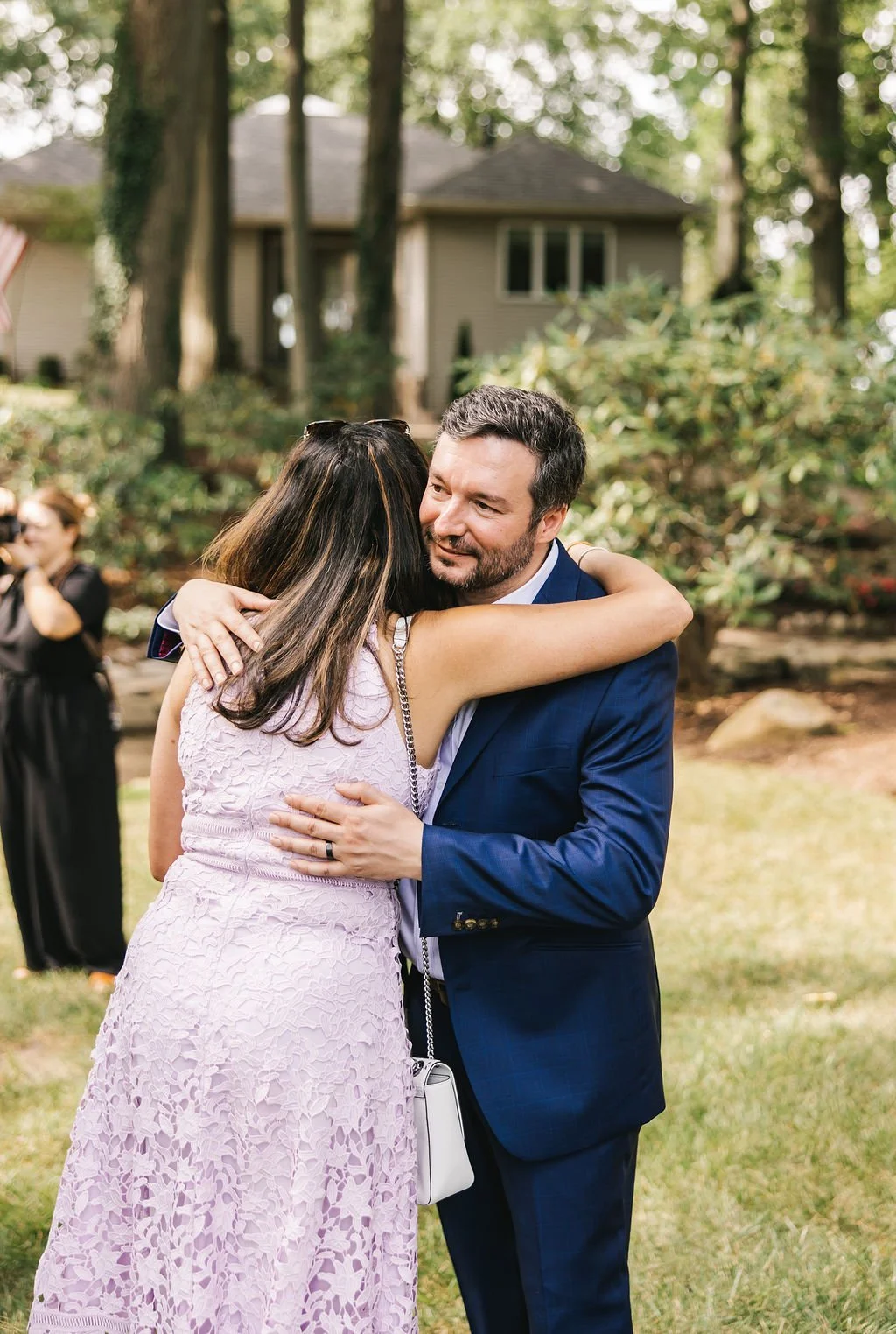 A man and woman hugging outdoors at a social gathering, with a house and trees in the background.