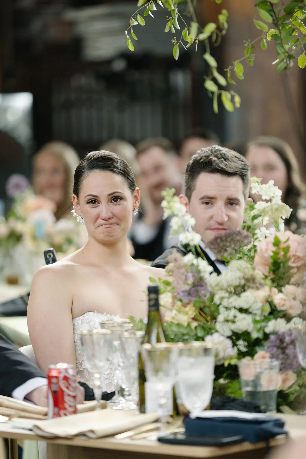 A bride and groom at a wedding reception, sitting at a table with drinks and flowers, with guests in the background, one of whom is crying.