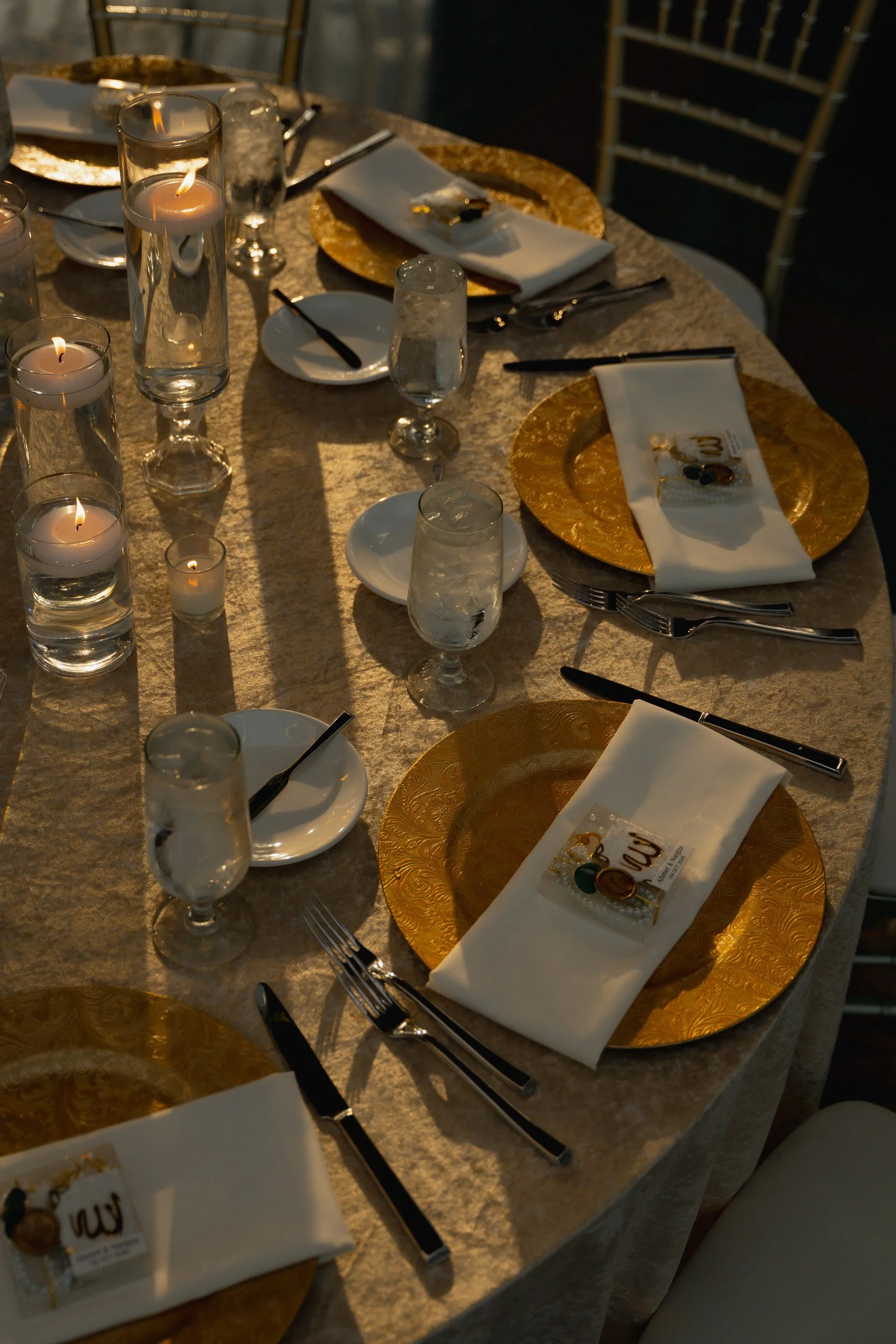 Elegant dining table set with gold chargers, white napkins, small favor boxes, glassware, and lit candles in a warm, ambient setting.