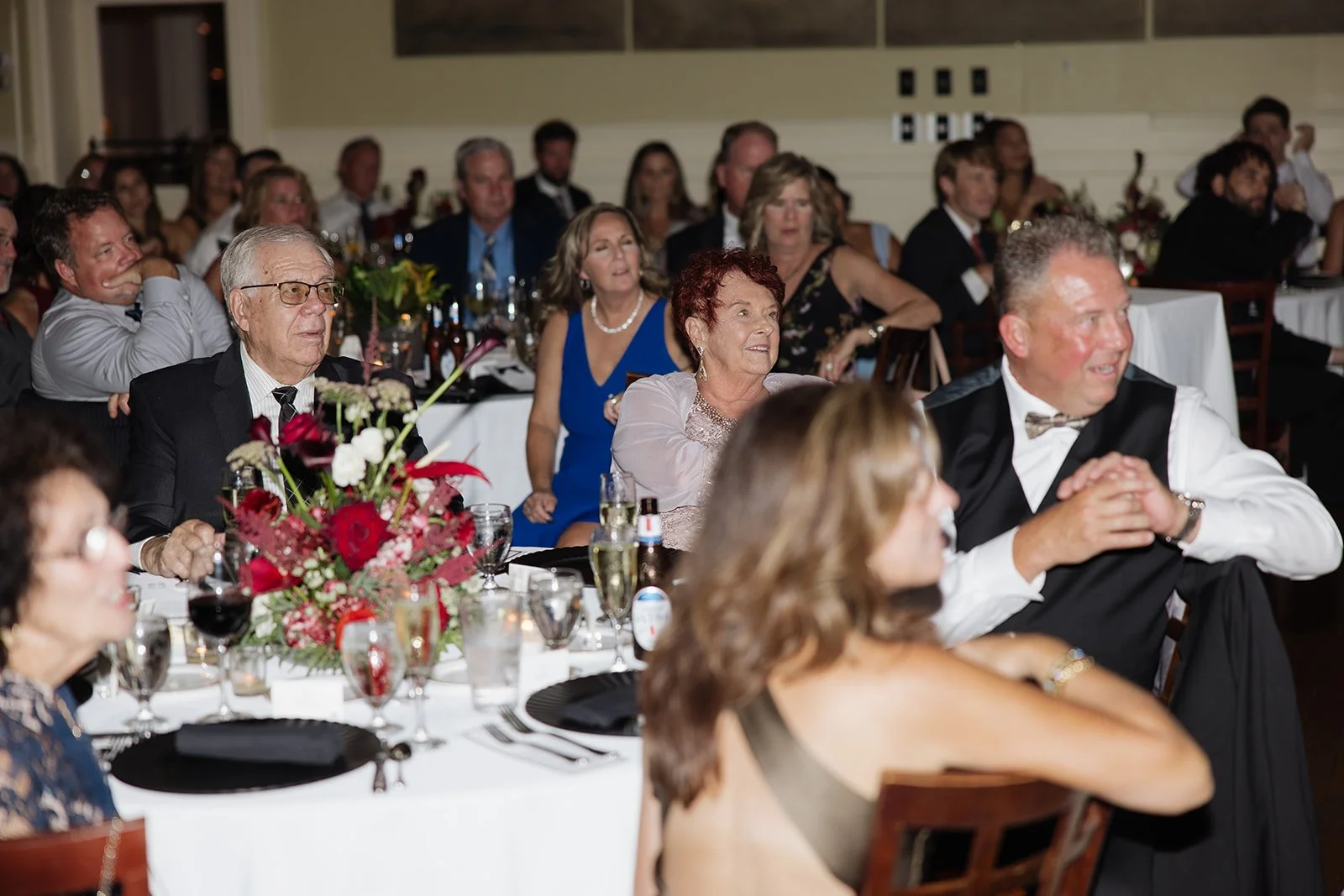 People sitting at round tables with white tablecloths and floral centerpieces, dressed in formal attire, at a banquet or celebration event.