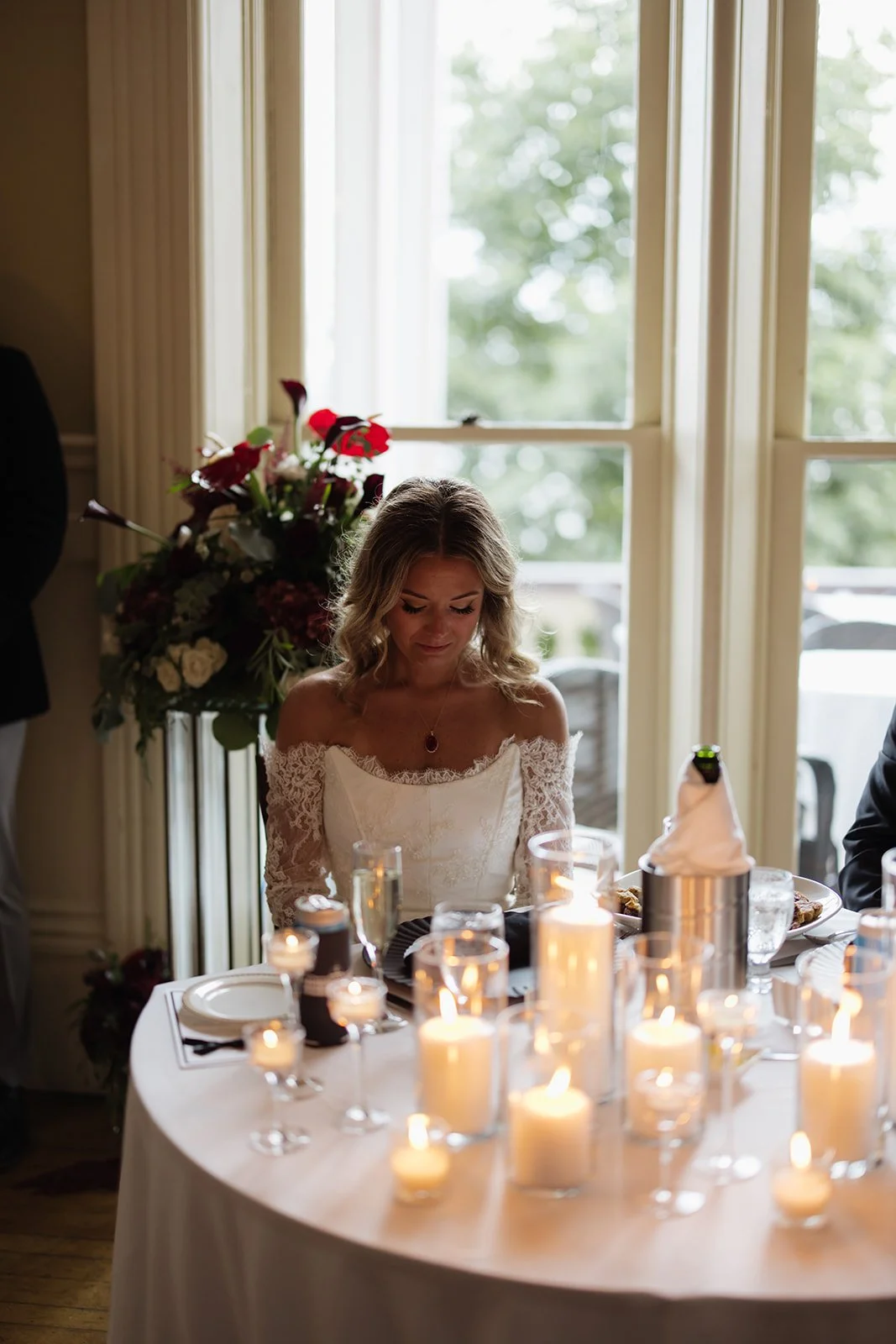 A woman in a white lace off-shoulder dress sits at a table with lit candles and glasses, inside a room near a large window with trees outside.