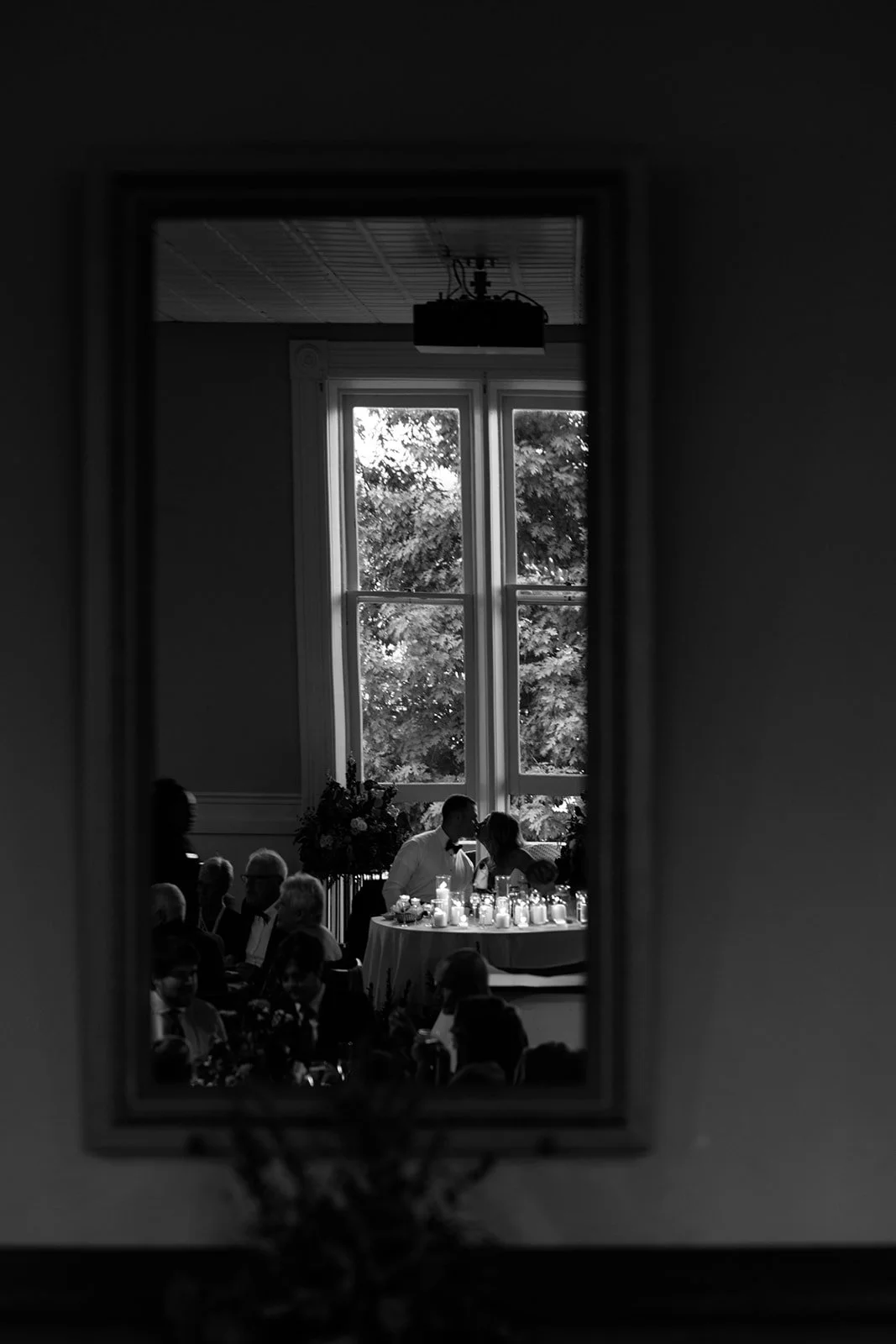 A mirror reflection showing a wedding reception with guests sitting at tables, a couple sharing a moment near a decorated table, large windows with trees outside, and candles on the tables.