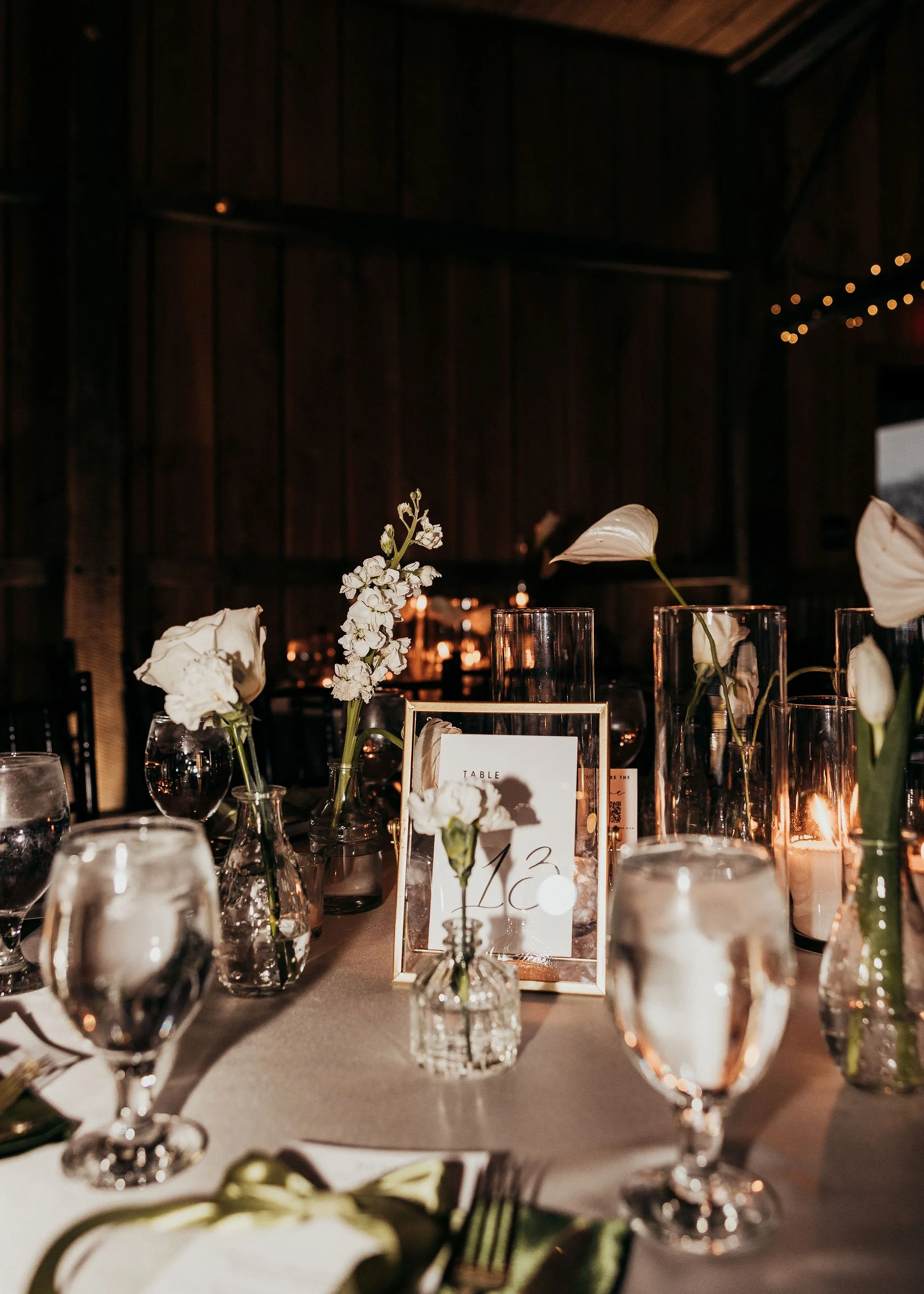 Table setting with white flowers in vases, glassware, candles, and a table number frame, in a dimly lit ambiance with wooden walls and string lights.