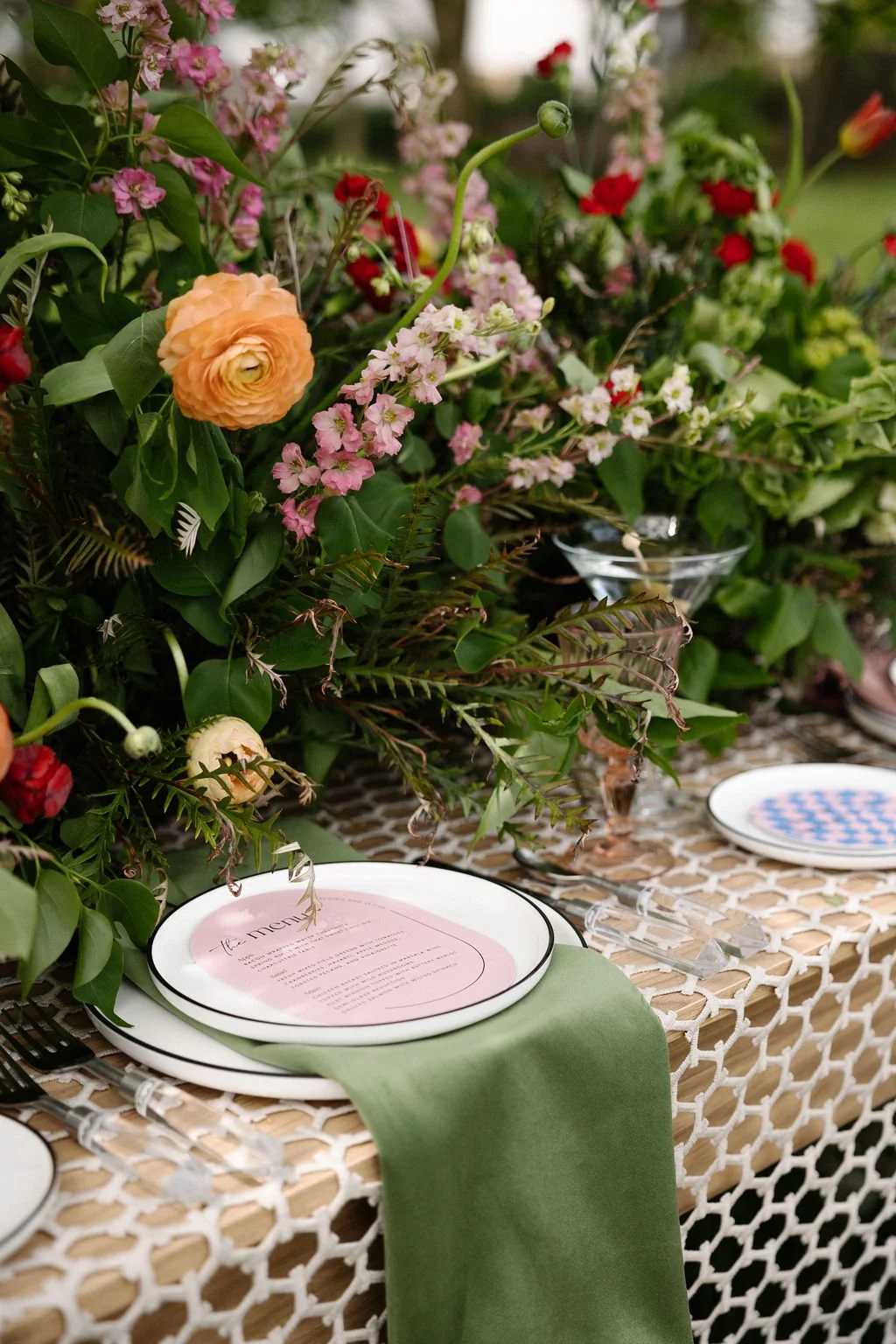 A table decorated with a lush arrangement of pink, peach, and red flowers and greenery, set with white plates, clear glassware, and black utensils, on a woven tablecloth with a green table runner.