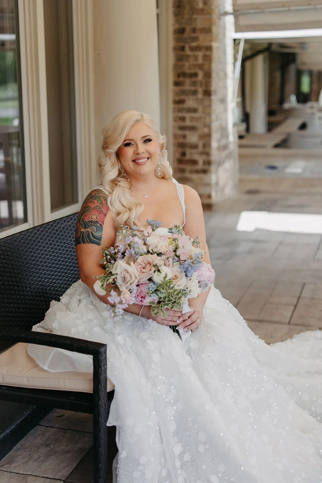 A smiling bride with blonde wavy hair, wearing a white wedding dress, sitting on a patio bench, holding a large bouquet of pink, purple, and white flowers, with a tattoo sleeve on her left arm.