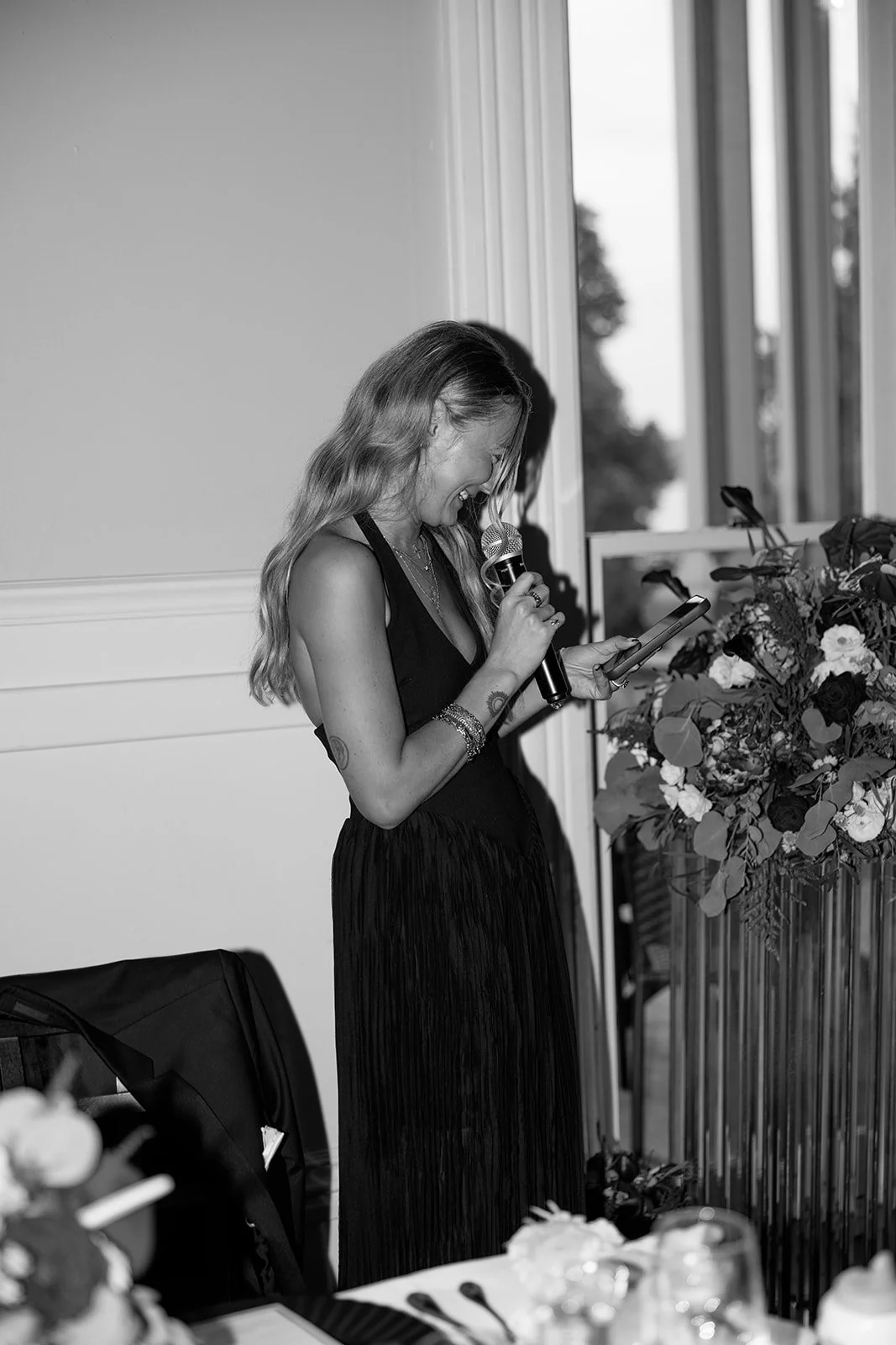 A woman with long hair, wearing a sleeveless dress, speaking into a microphone while looking at her phone near a large floral arrangement, indoor setting with window in background.