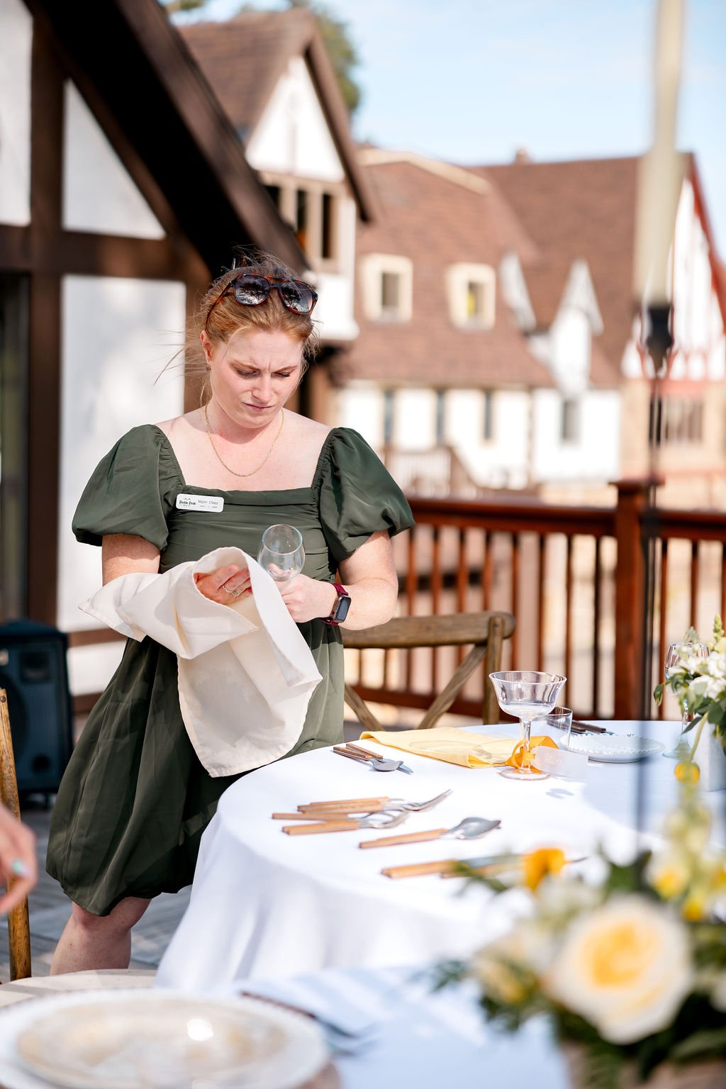 A woman with red hair, sunglasses on her head, and a dark green dress looks down at a wine glass in her hand while holding a napkin, standing next to a table set with plates, utensils, and glasses outdoors in front of houses.