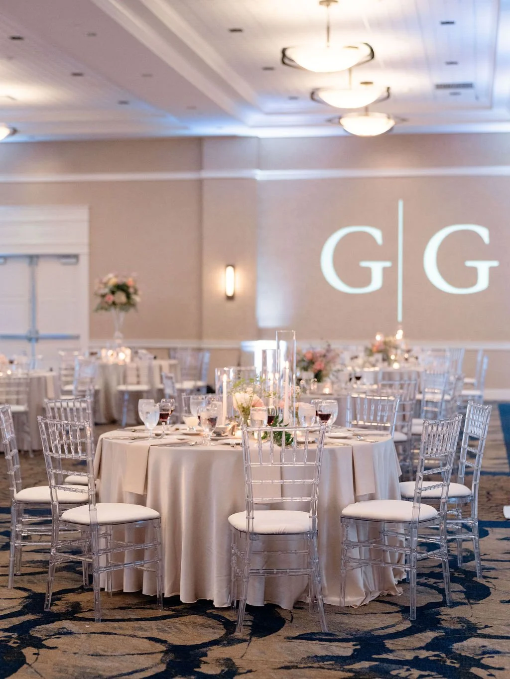 Elegant banquet hall with round tables draped in beige tablecloths, decorated with floral centerpieces and tall glass candle holders, surrounded by clear acrylic chairs. A large projection on the wall displays the initials G | G.